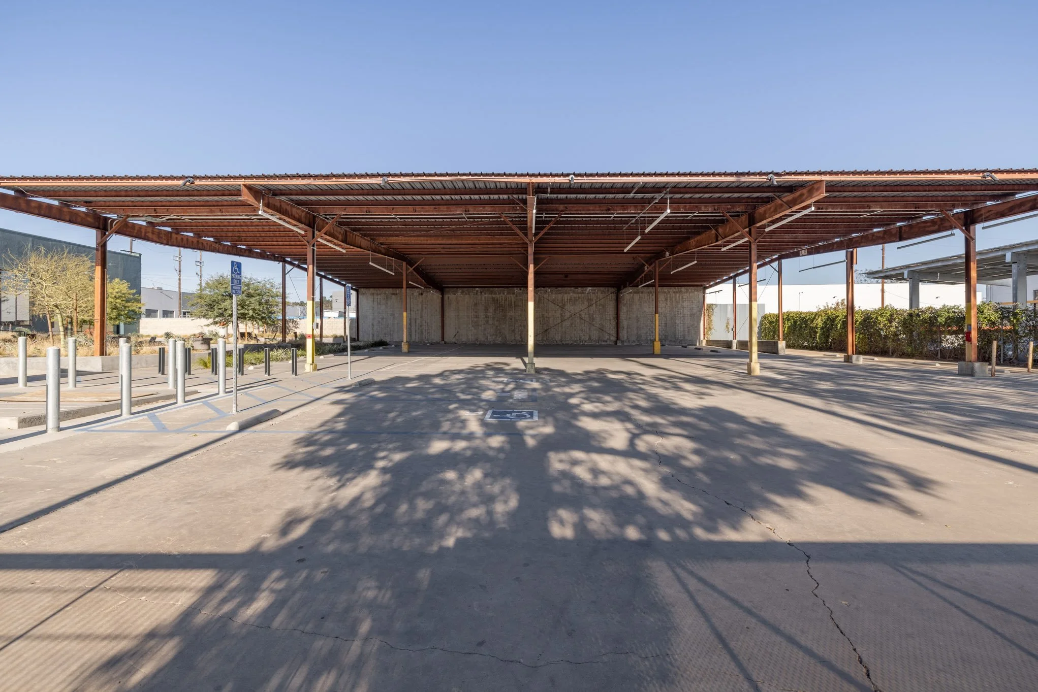 Empty outdoor parking lot with a large wooden roof structure, a designated accessible parking spot, and shadows of trees on the pavement.