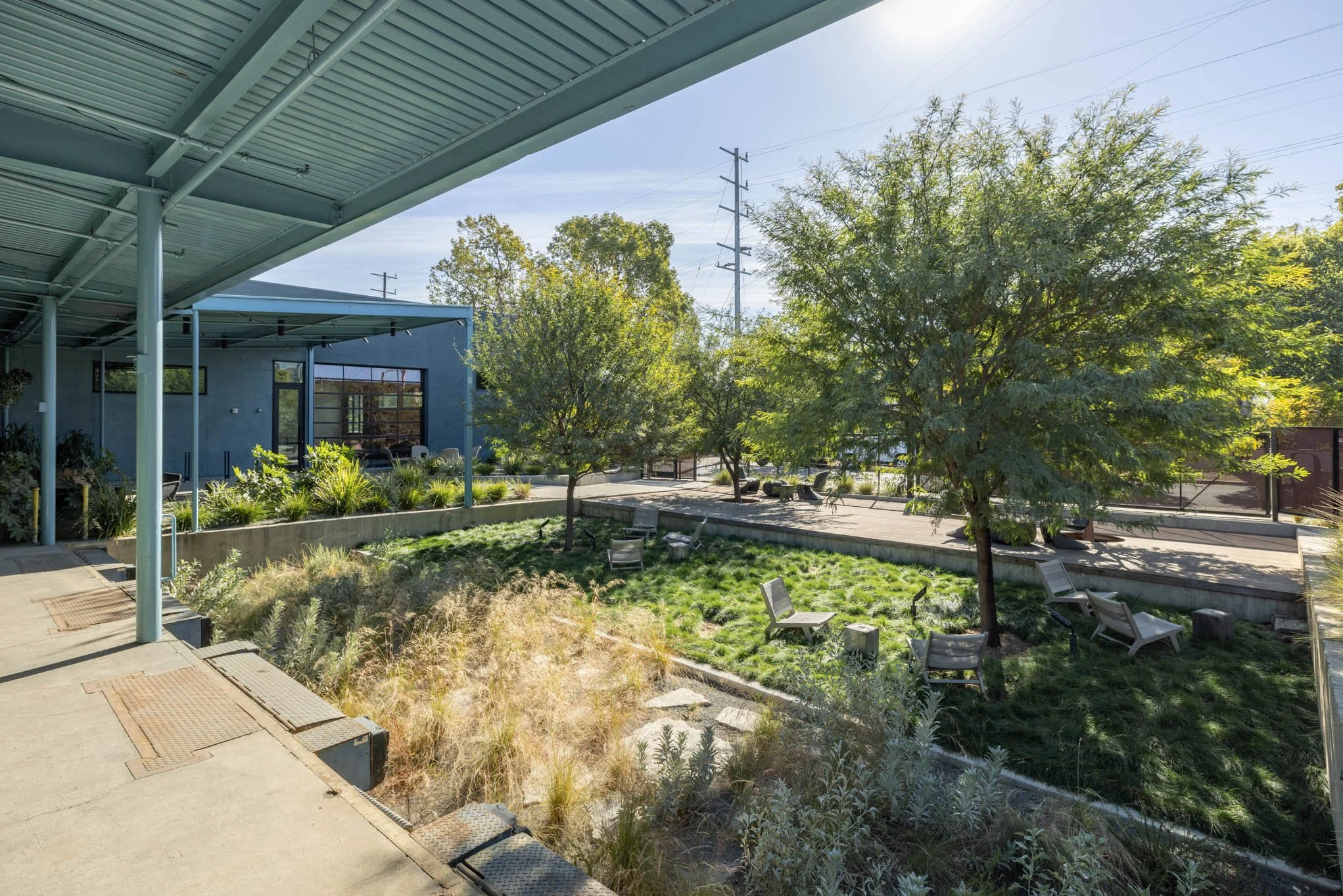 Outdoor courtyard with trees, shrubs, benches, and a building with large windows, under a covered walkway with a cloudy sky.
