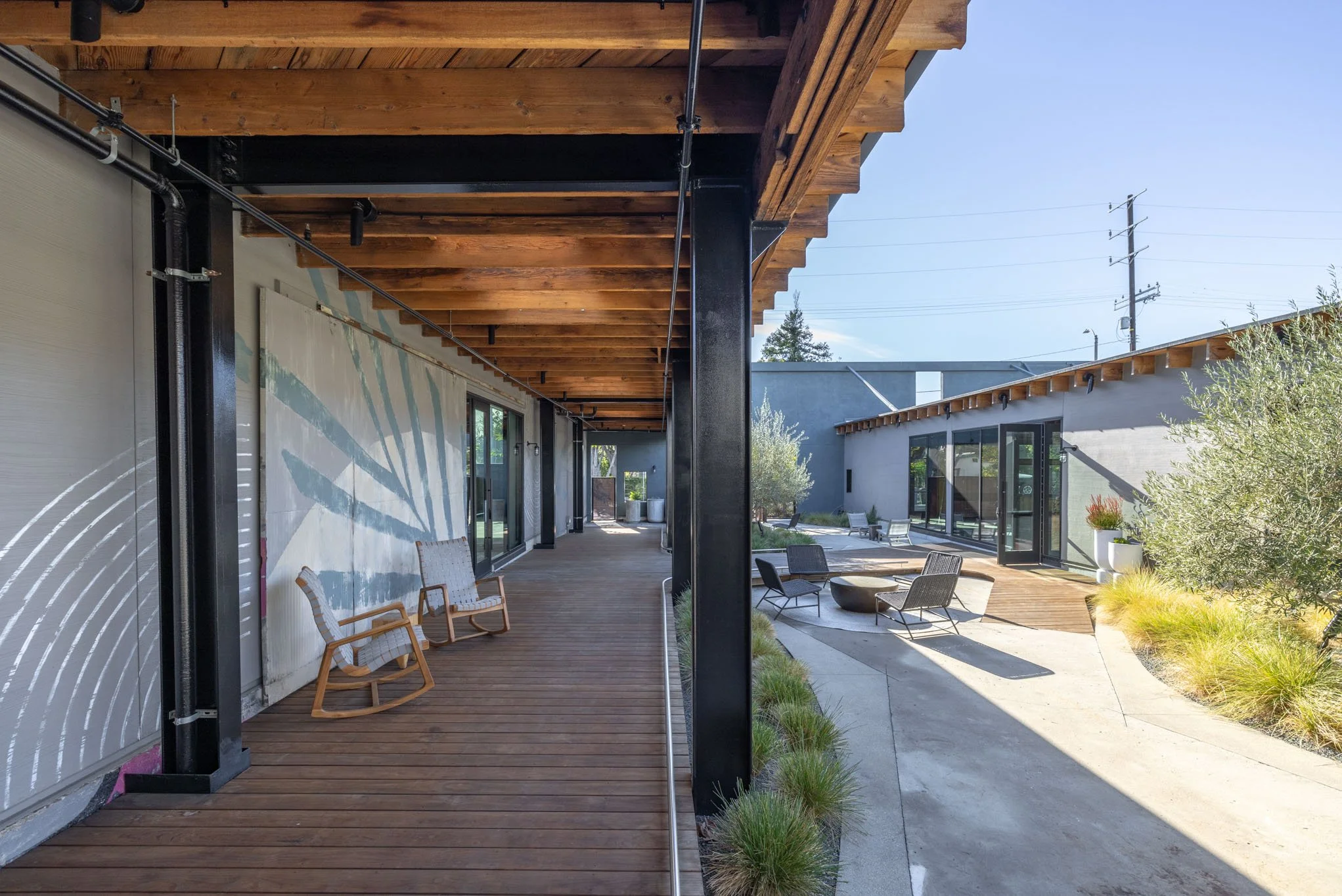 Outdoor patio with wooden deck, modern chairs, large sliding glass doors, and landscaped garden area with plants and concrete pathway.
