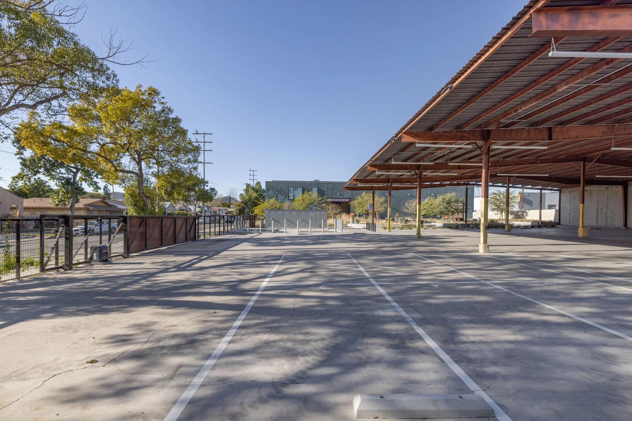 Empty outdoor parking lot with marked parking spaces, a covered area on the right, trees on the left, and a building in the background under a clear blue sky.