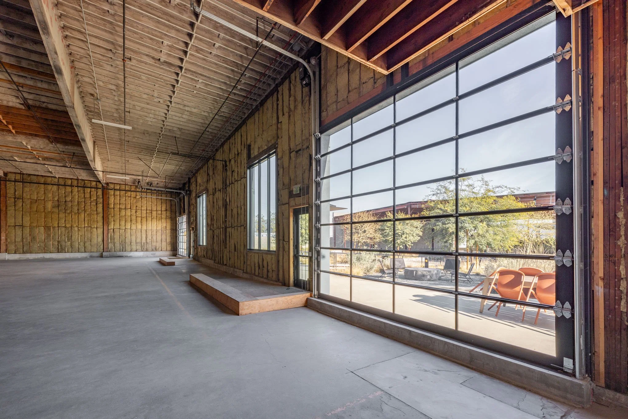 Inside an unfinished building with large garage-style windows, exposed ceiling beams, and insulation on the walls, with a concrete floor and outdoor patio area visible through the windows.