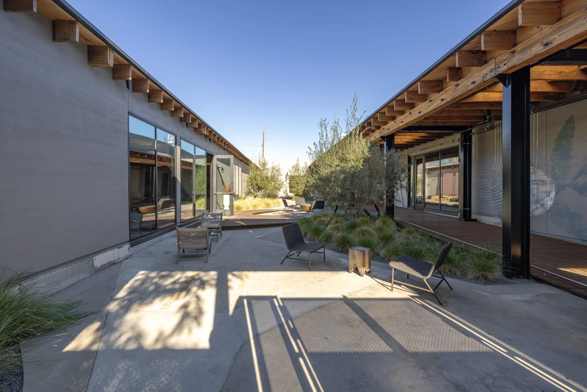Modern outdoor courtyard between two buildings with glass walls, patio chairs, and landscaped greenery under a clear blue sky.