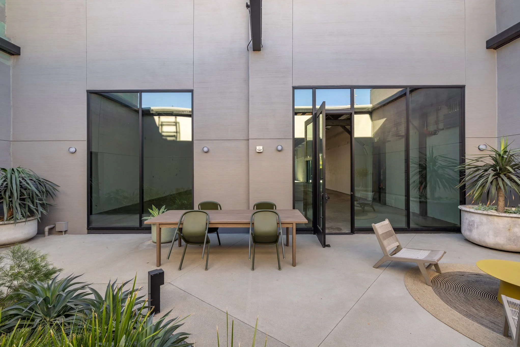 Modern outdoor patio with a rectangular wooden table, six green chairs, lounge seating, and large planters with greenery in front of a building with large glass doors and windows.
