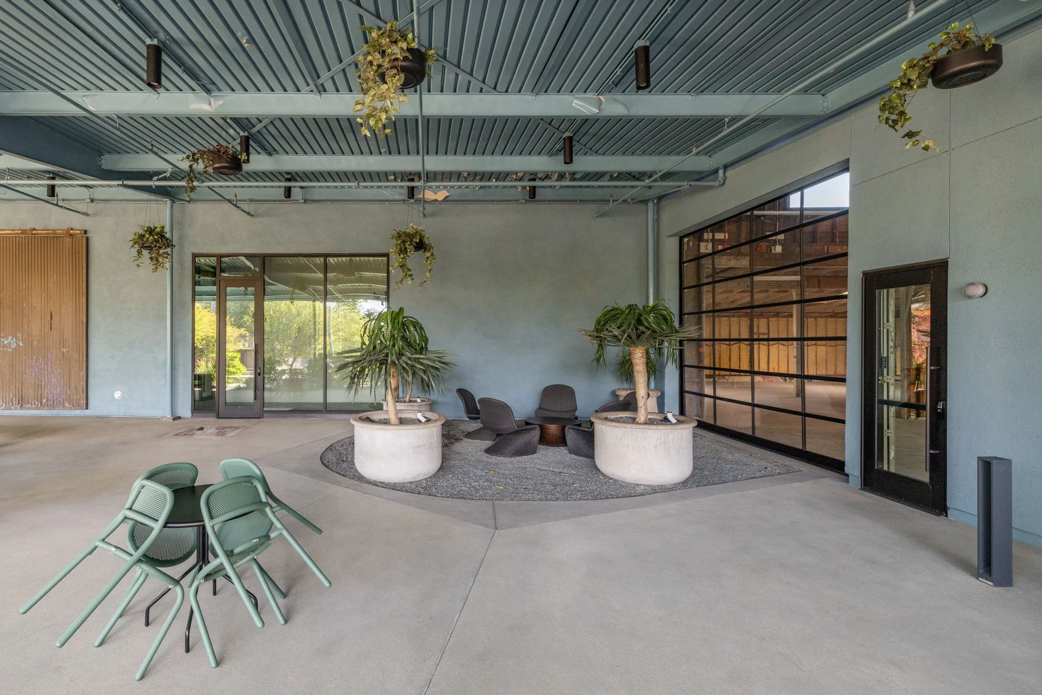 An indoor seating area with black chairs and two large potted plants, green chairs stacked on the floor, glass doors, and hanging plants from the ceiling.