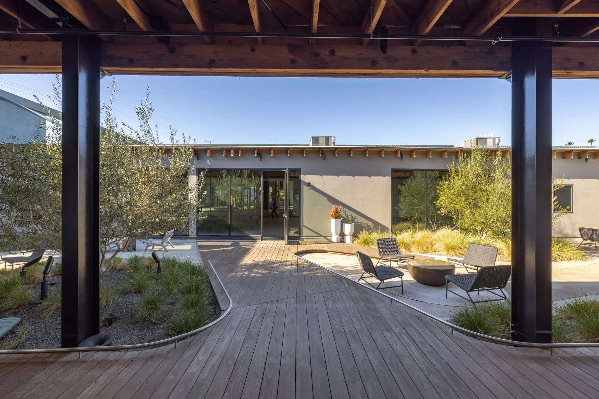 Modern outdoor patio with wooden flooring, outdoor seating, potted plants, and a view of a single-story building with large glass doors and windows, under a wooden roof