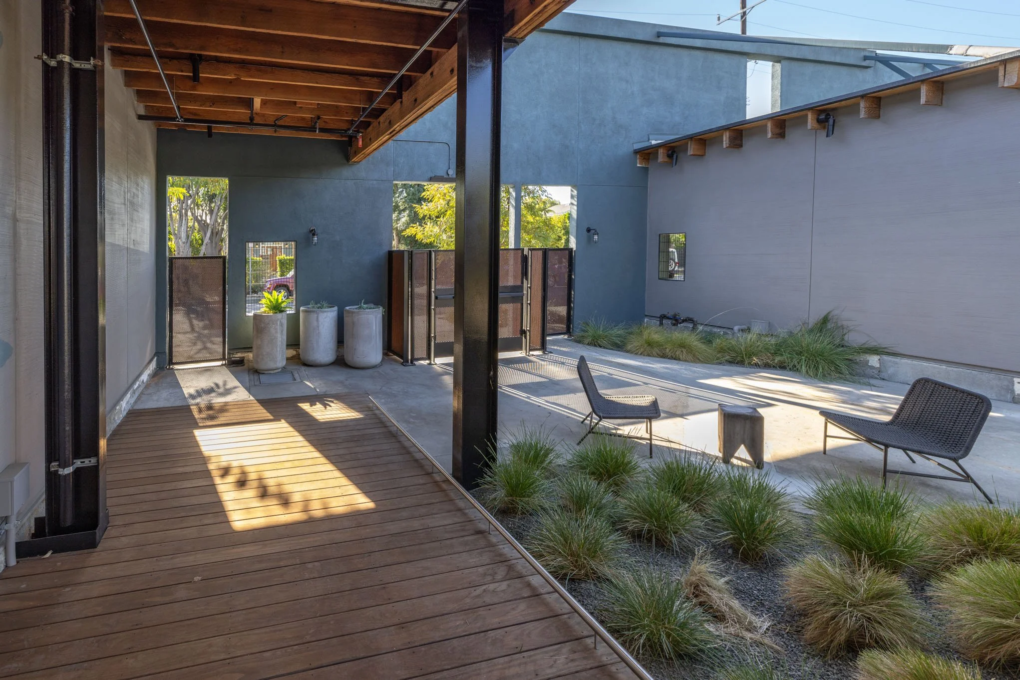 An outdoor patio area with a wooden deck next to a concrete area with chairs, modern planters with plants, and surrounding grass and shrubs, enclosed by dark-colored walls and screens.