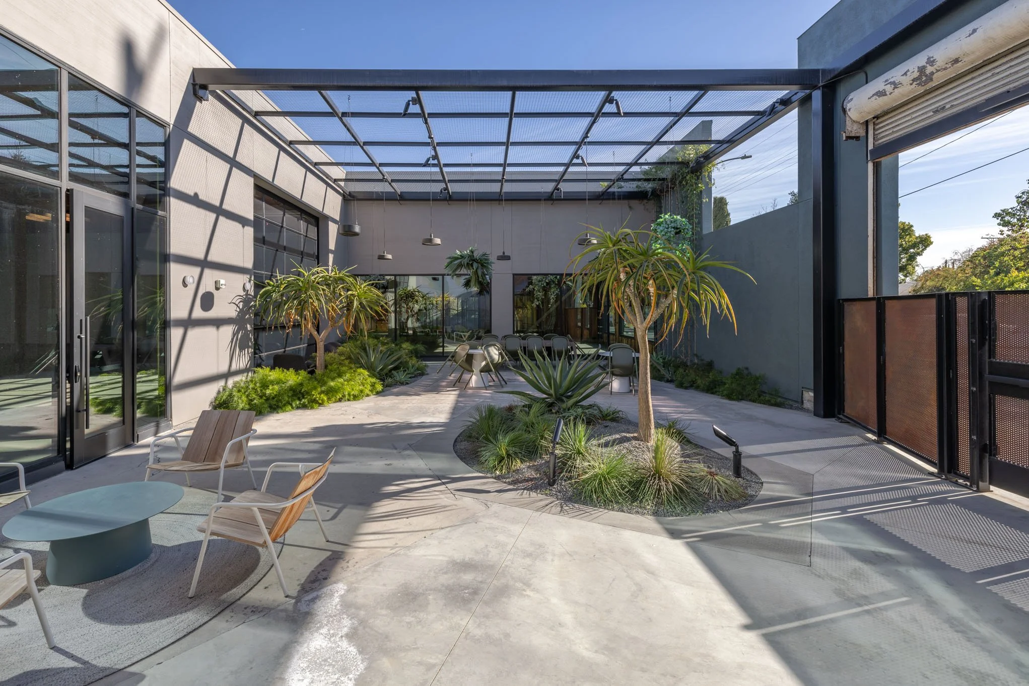 Modern outdoor patio with greenery, lounge chairs, round tables, and metal chairs under a glass pergola, adjacent to a building with large windows and an industrial-style ceiling.