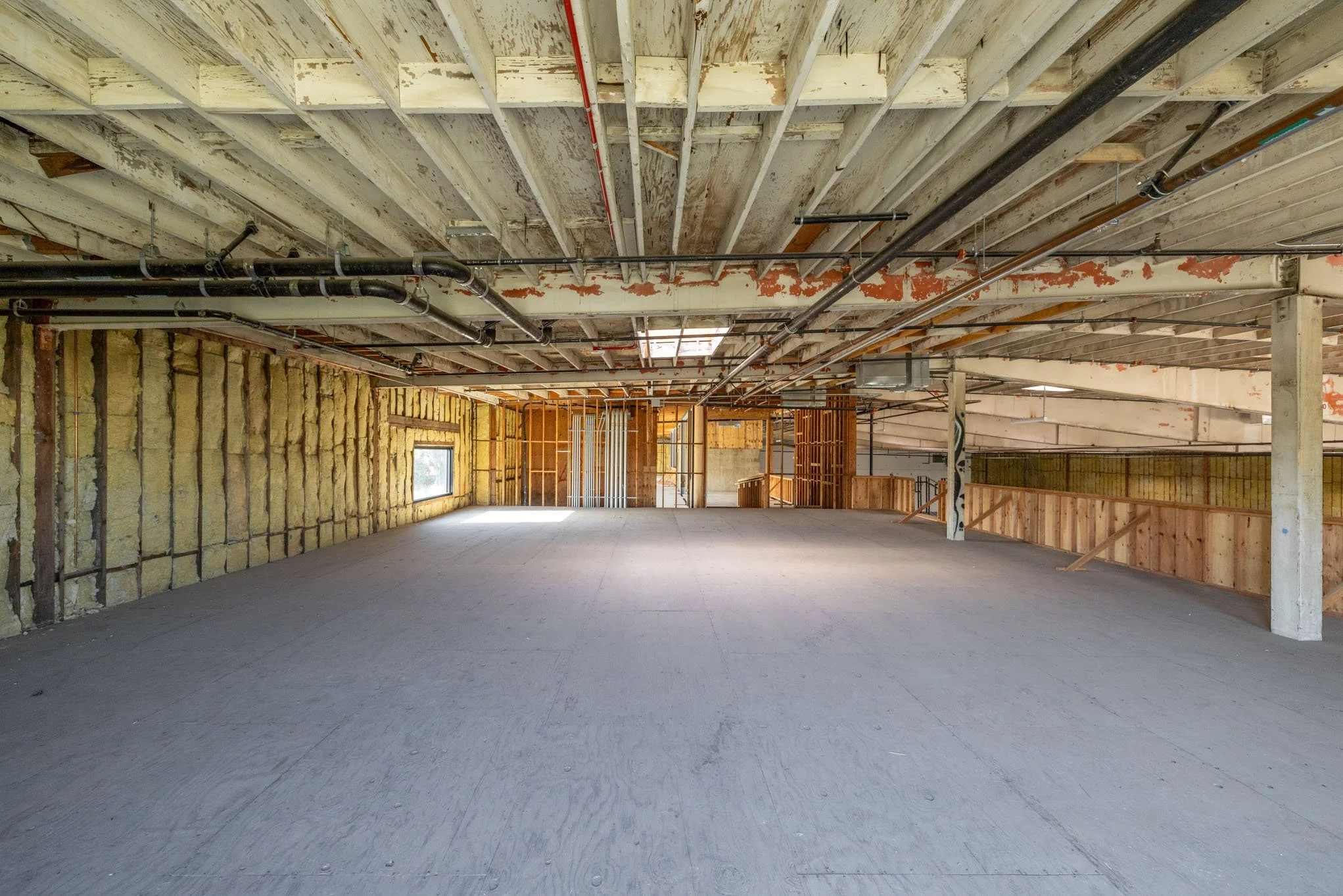 Interior view of a building under construction with exposed ceiling beams, insulation on walls, and a concrete floor.