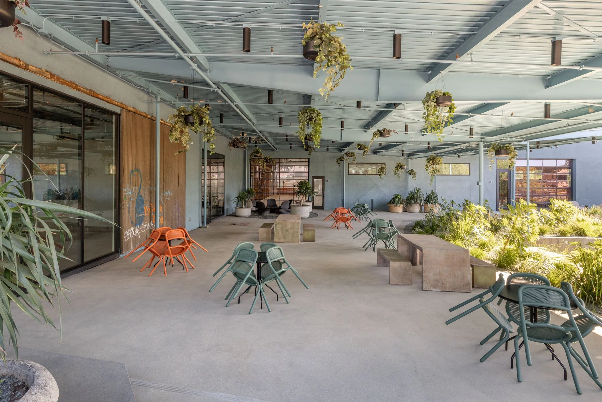 An outdoor patio area with multiple green and orange chairs, concrete tables, and lush green plants, under a blue metal roof with hanging plants.