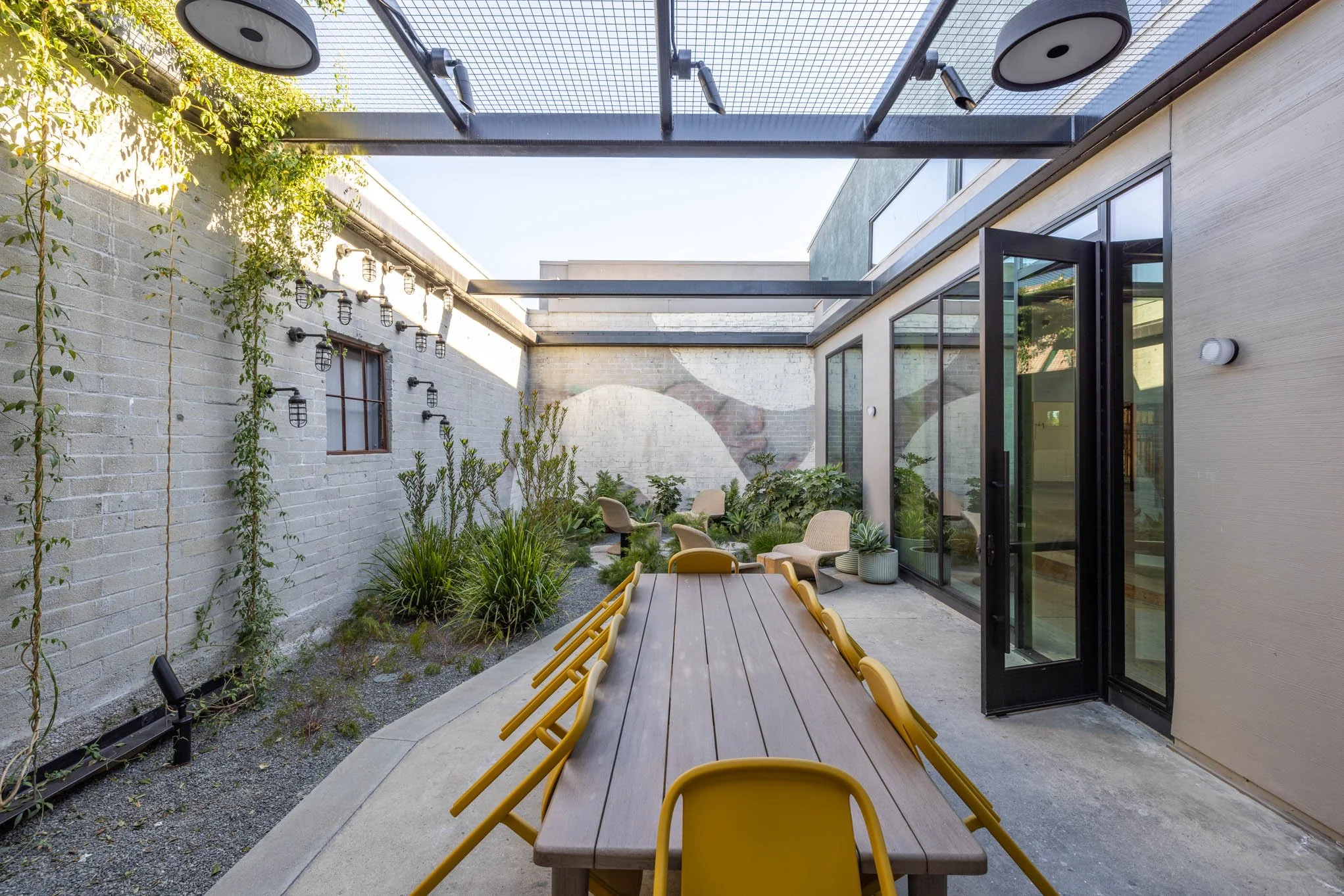 An outdoor patio area with a long wooden dining table surrounded by yellow chairs, a small sitting area with beige chairs and potted plants, enclosed by a brick wall and a beige wall with a glass door, and shaded by a pergola with black hanging lamps