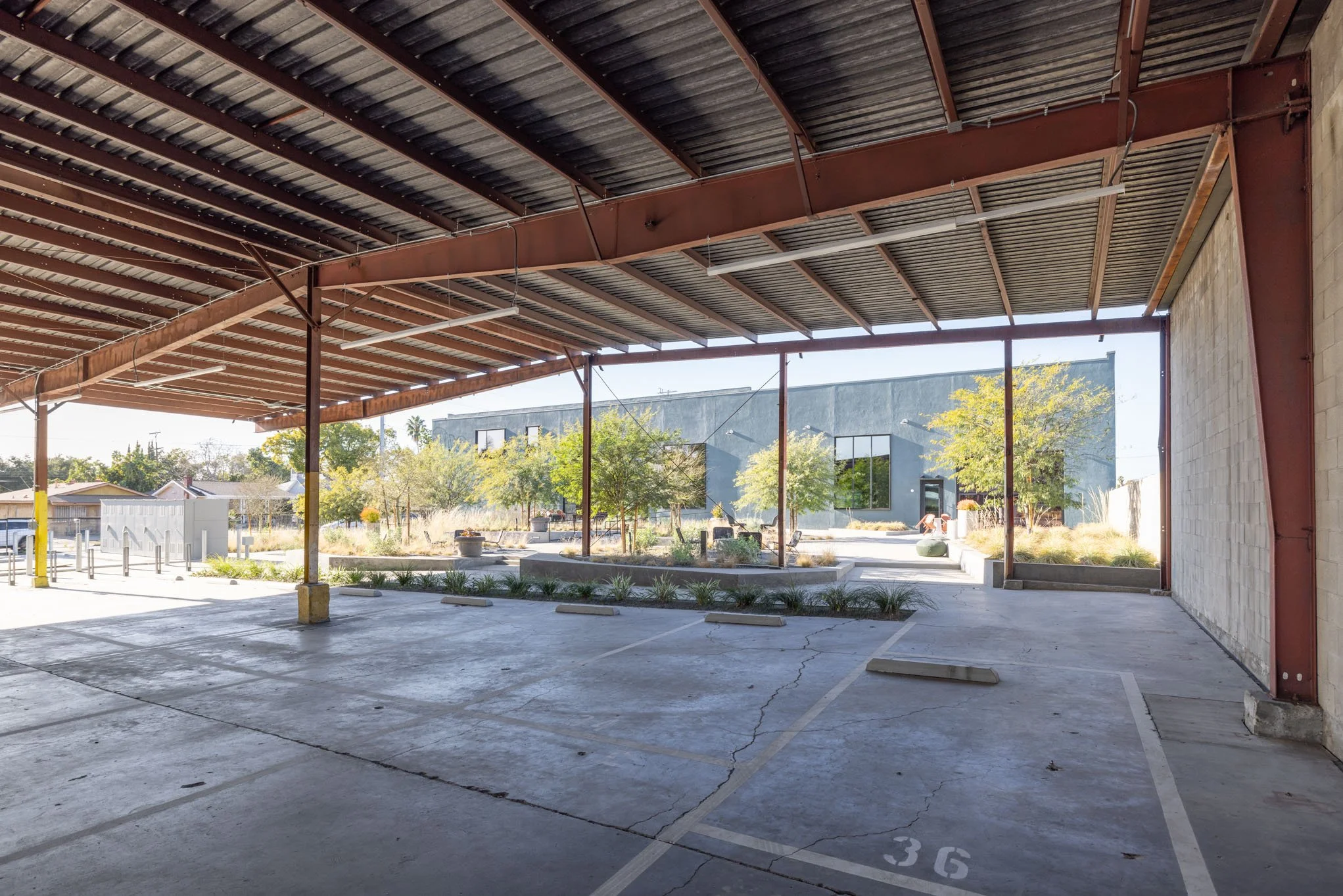 Empty parking lot under a metal overhang with a view of trees and a modern gray building in the background.