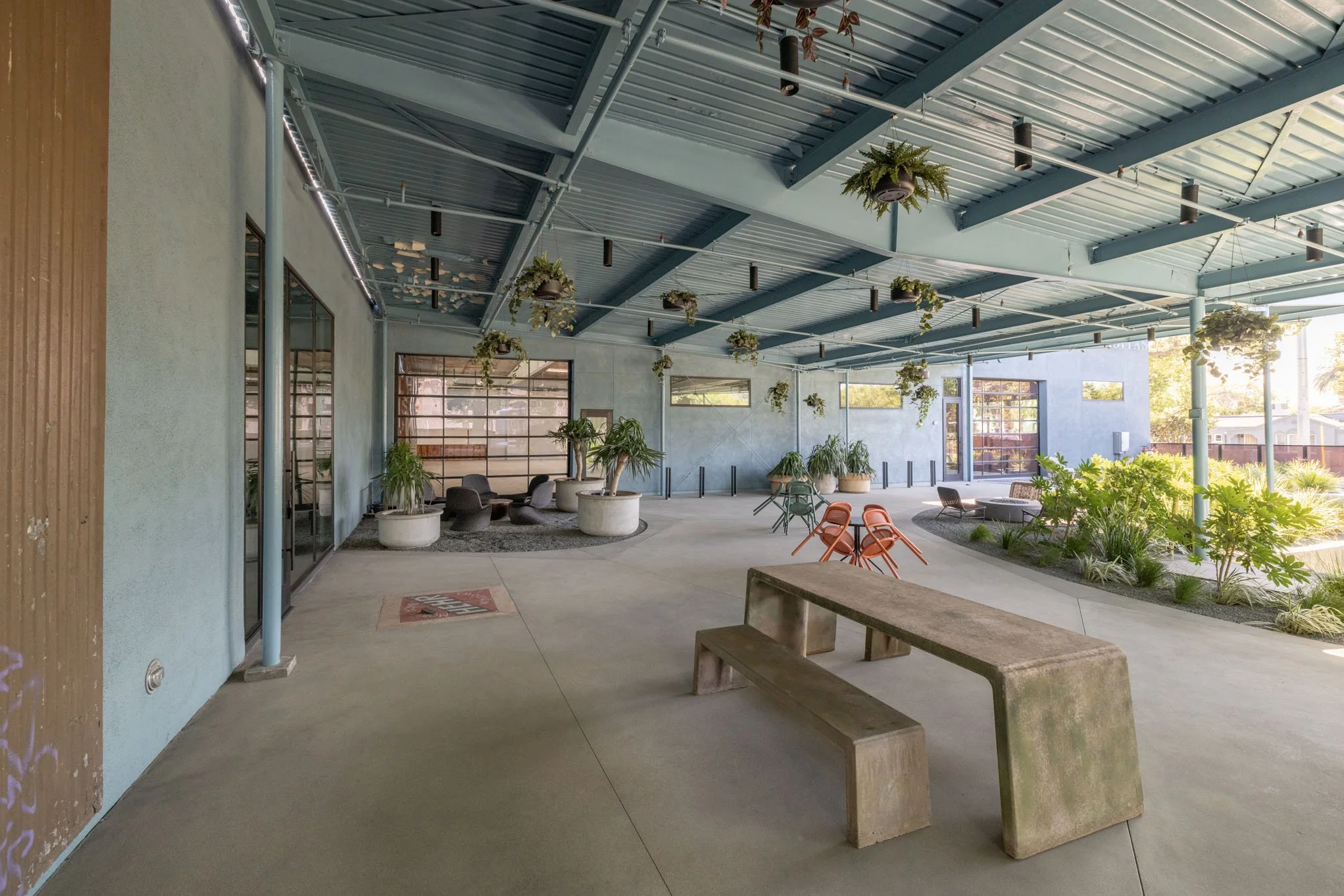An outdoor patio space with concrete flooring, potted plants, hanging plants from the blue metal ceiling, outdoor seating, and landscaped garden areas, illuminated by natural sunlight.