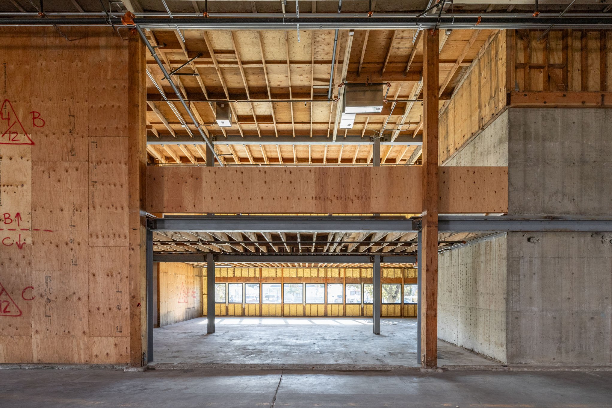 Interior view of an under-construction building with exposed wooden framing, concrete walls, and large windows at the far end.