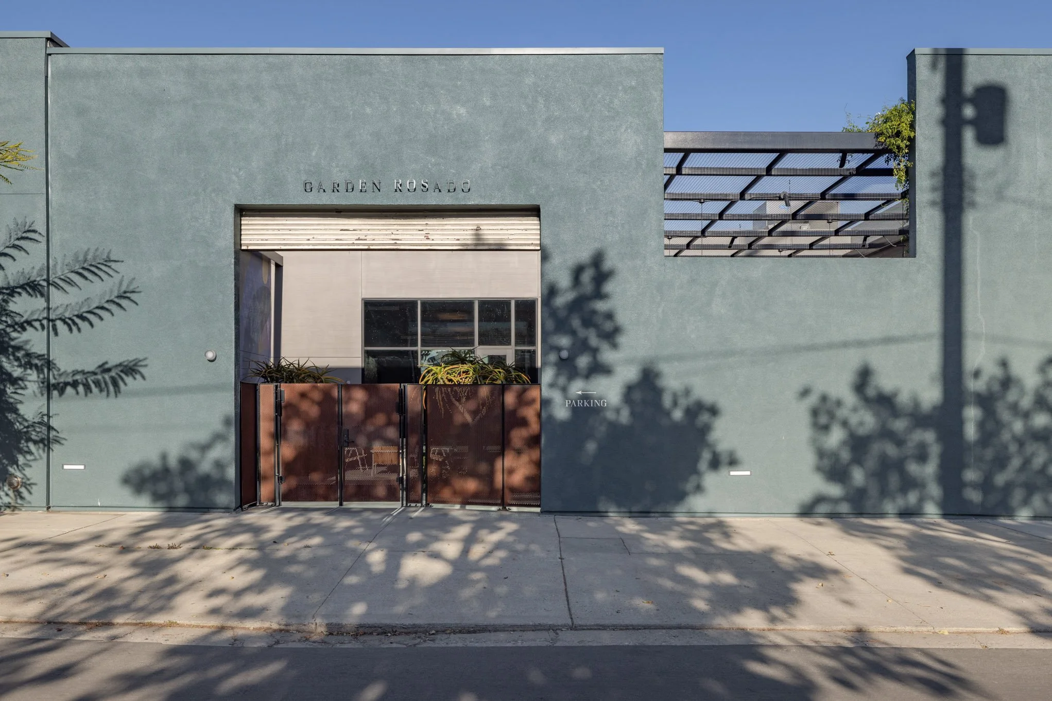 Modern building with a sign that reads 'GARDEN ROSADO,' a glass window, a metal gate, and a small parking area, casting shadows on the sidewalk.