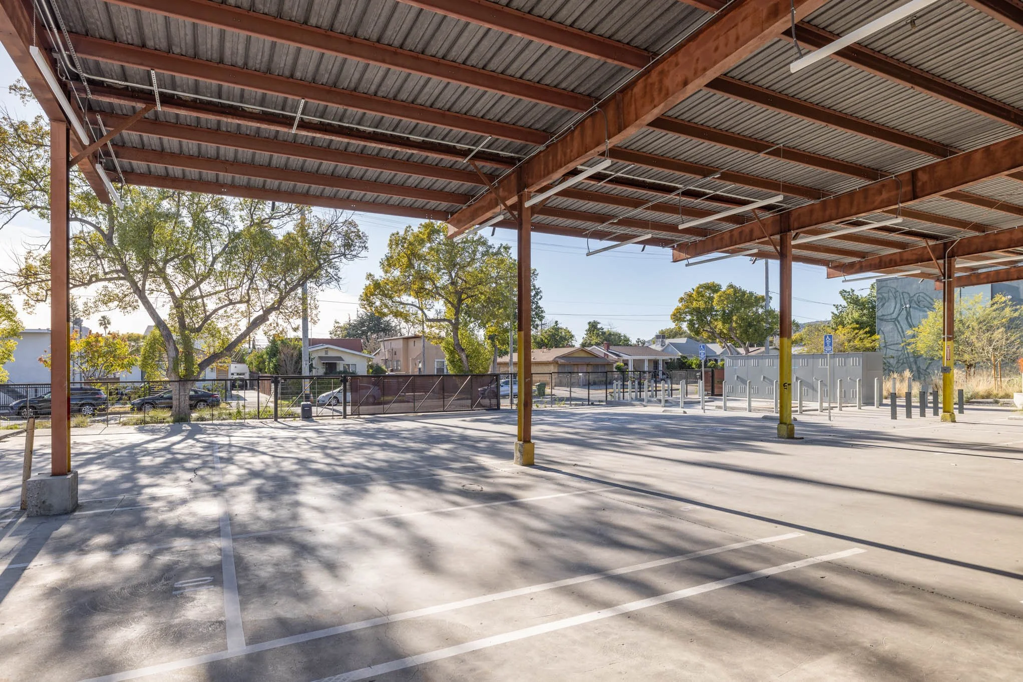 Empty parking lot under a metal roof structure, with trees and residential houses in the background.
