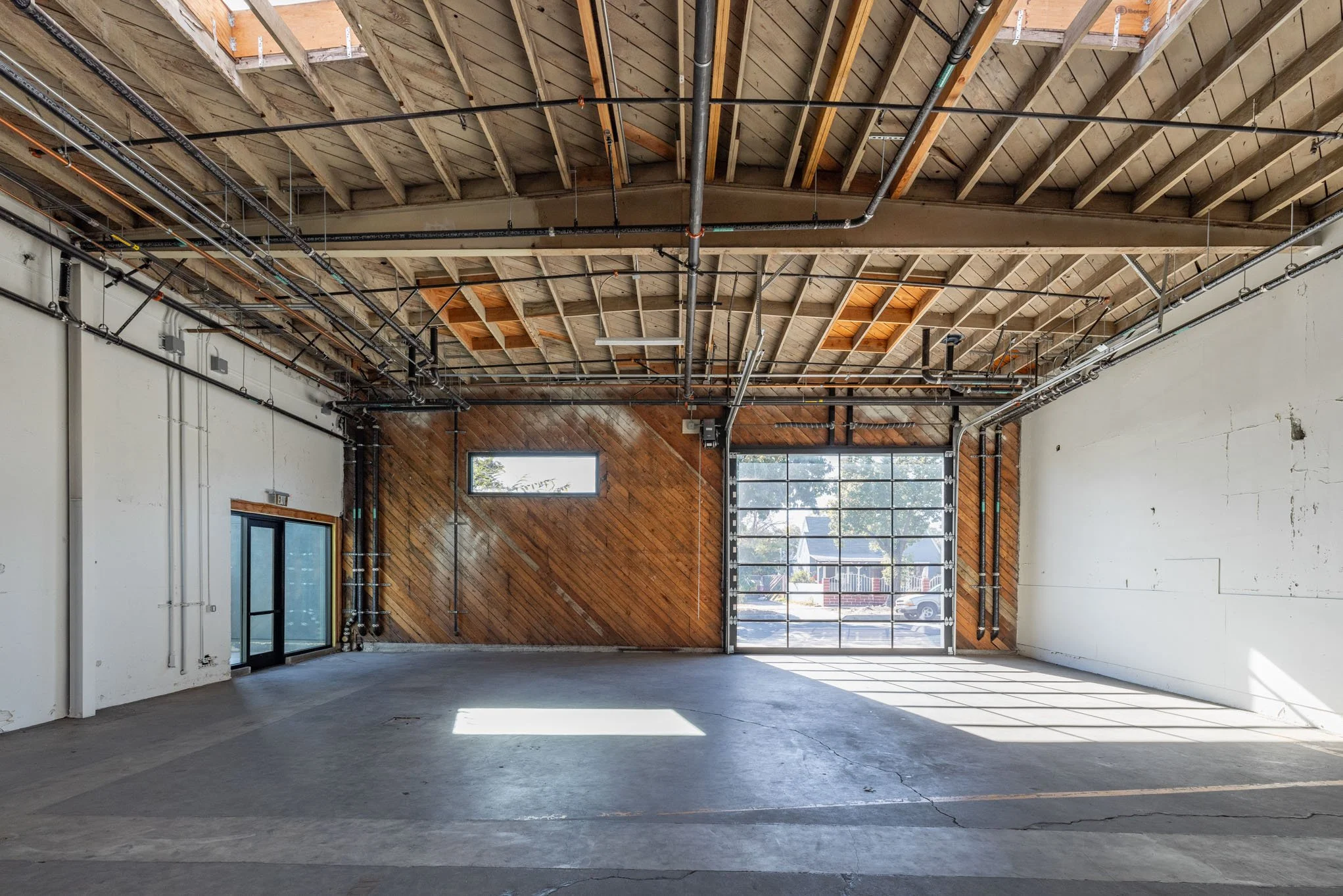 Empty industrial-style garage with a large glass roll-up door, wooden wall paneling, and exposed ceiling pipes.
