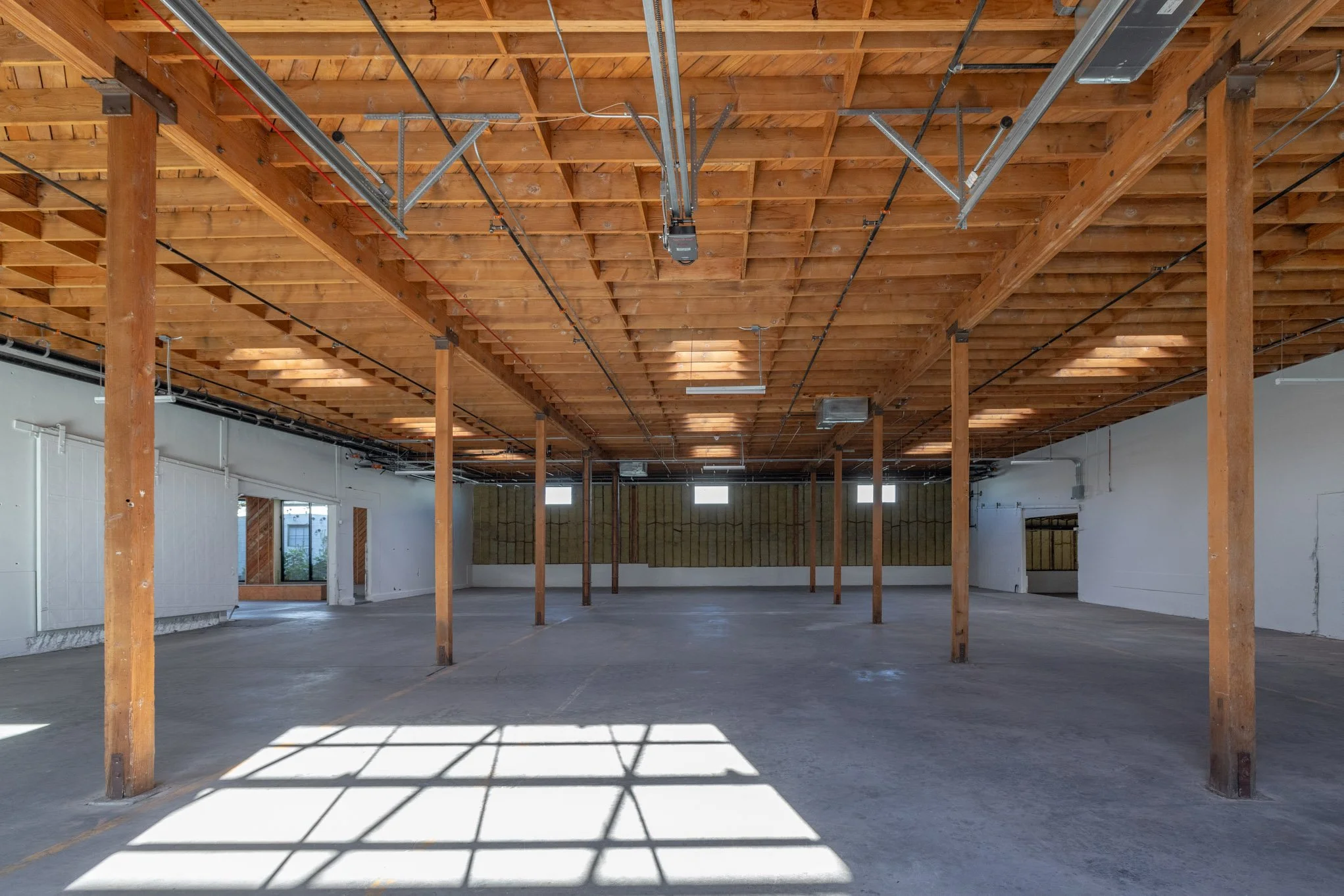 Empty indoor space with wooden support beams, unfinished ceiling, and concrete floor, illuminated by natural light through windows, indicating a building under construction or renovation.