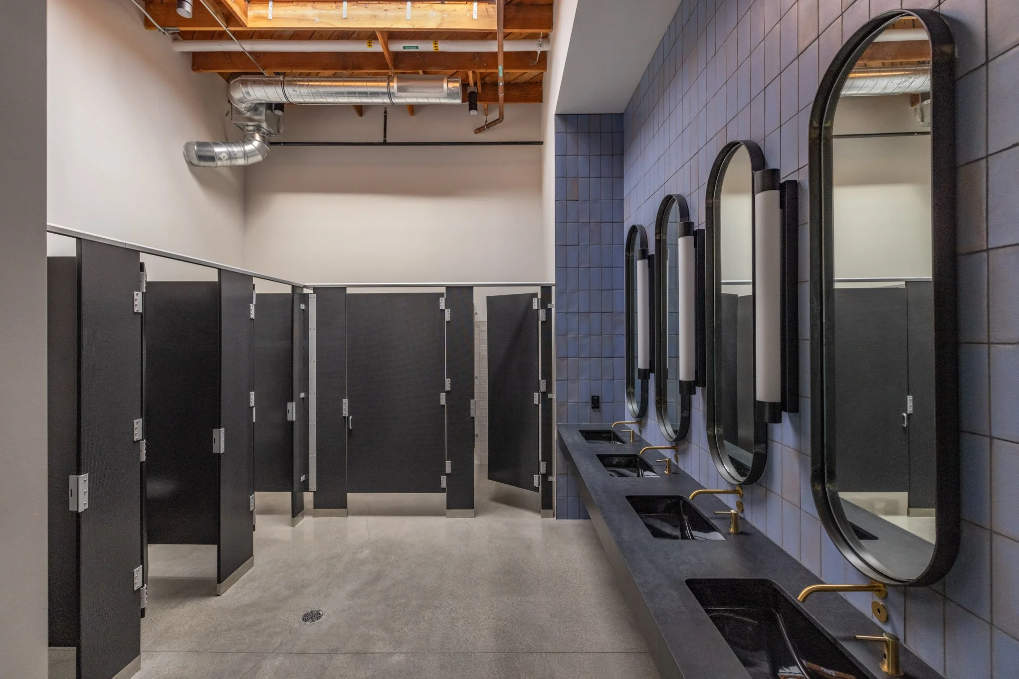 Public restroom with black lockers, a row of three mirrors with gold faucets, and blue tiled wall.