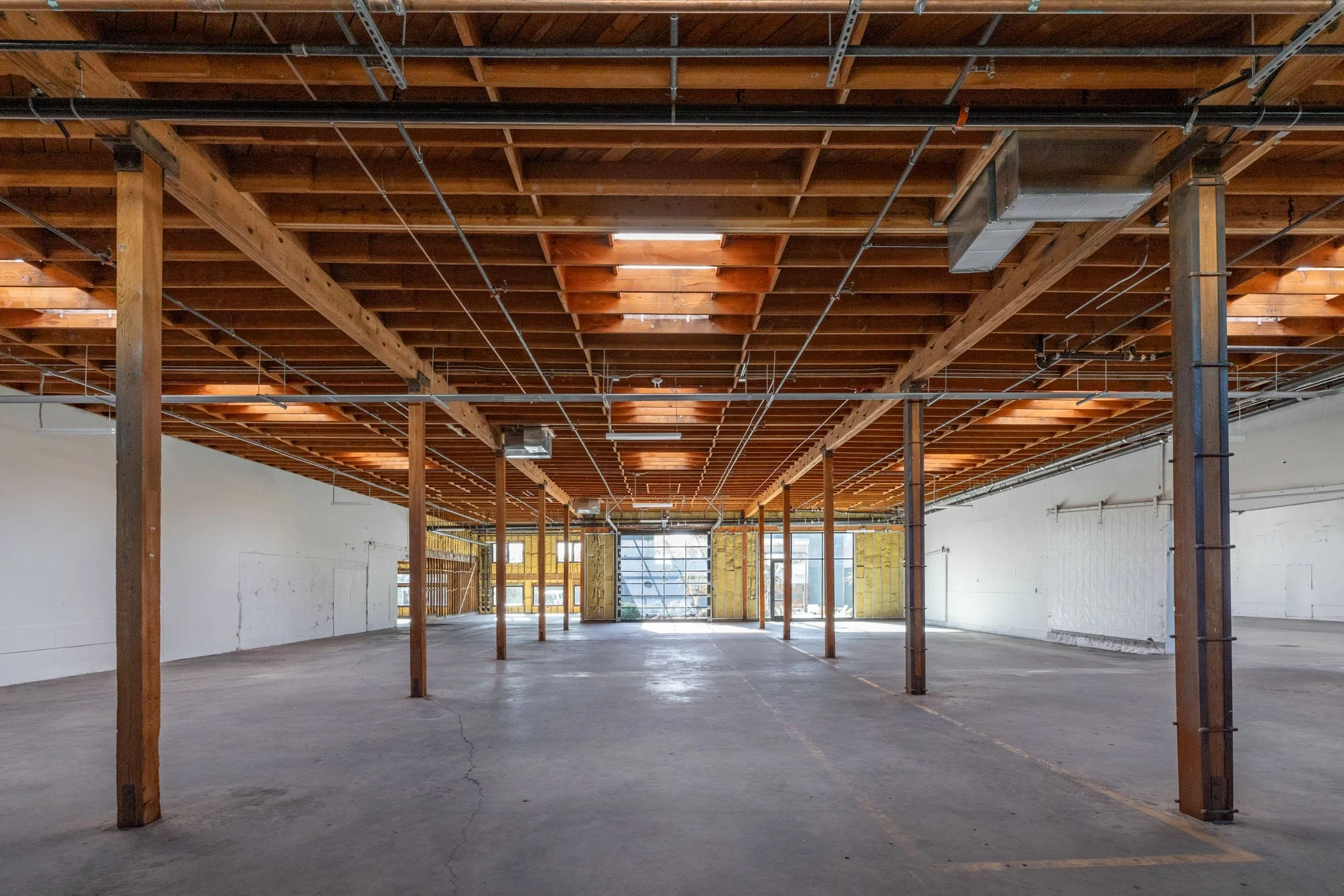 Empty building interior with exposed wooden ceiling beams, concrete floor, and large windows in the background.