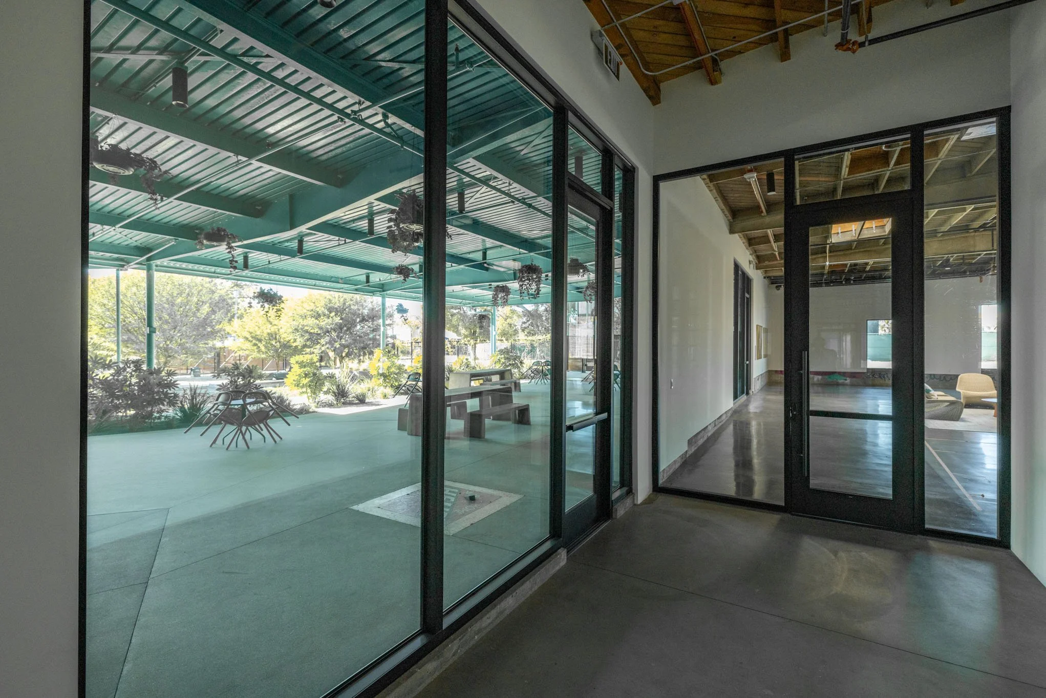 Interior view of a modern building with large glass windows, showing an outdoor covered patio with seating and greenery, and an adjacent room with wooden ceiling and minimal furniture.