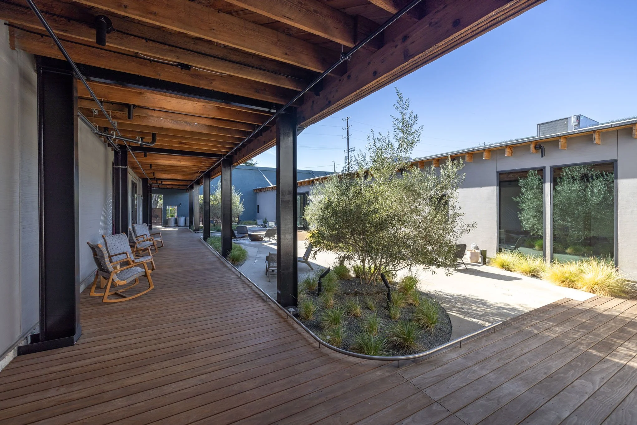 Outdoor patio with wooden deck, rocking chairs, and a landscaped garden area with shrubs and bushes, adjacent to modern buildings with large windows.