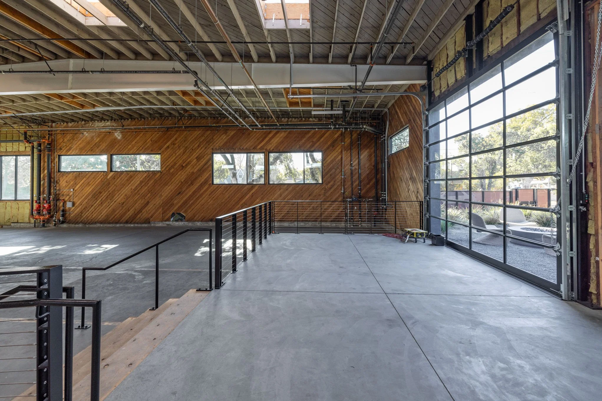 Interior of an unfinished industrial or commercial building with concrete floors, exposed ceiling beams, and large garage door with glass panels, bright natural light, and views of outdoor seating area.