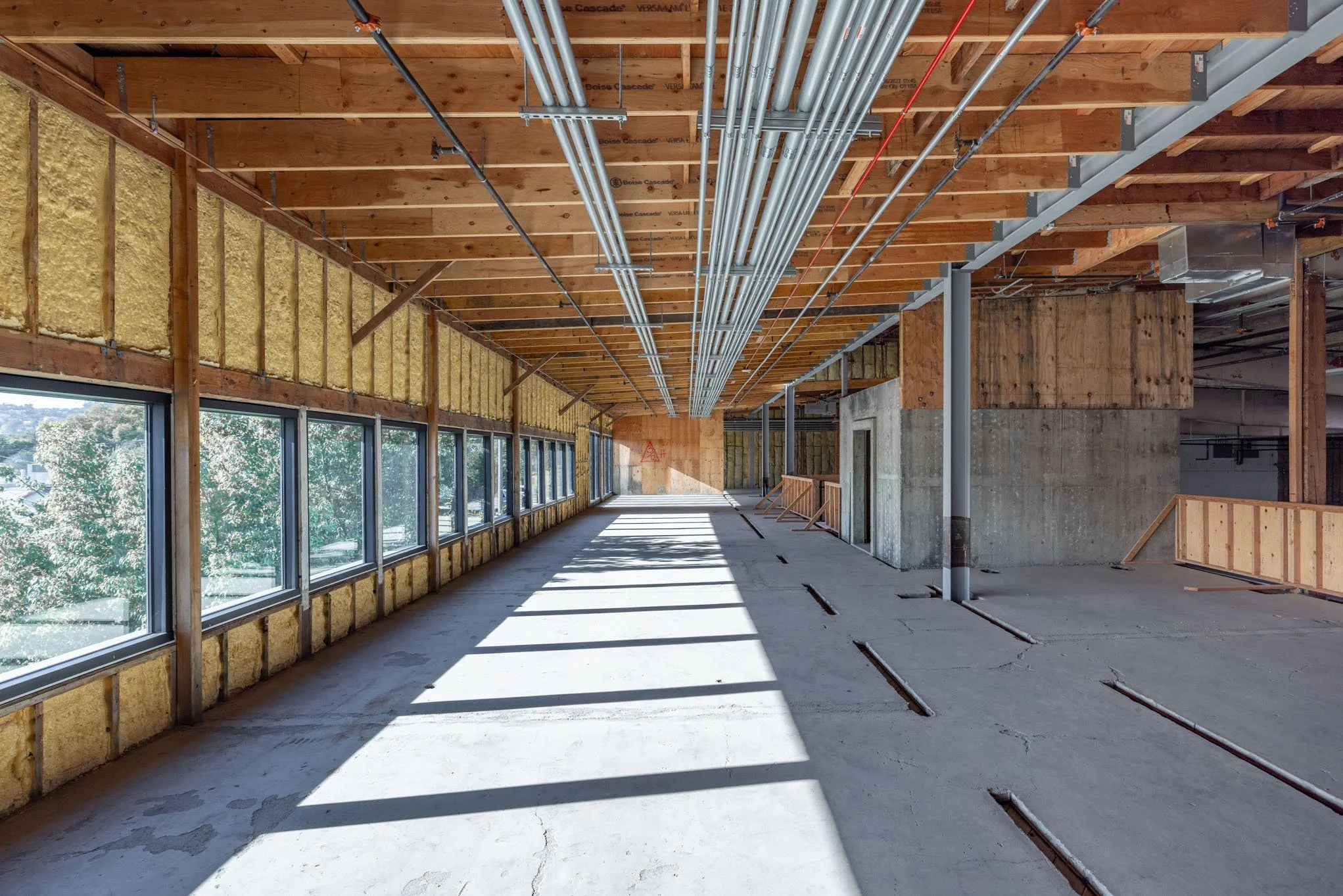 Unfinished indoor construction space with large windows, exposed wood beams, insulation, and pipes on the ceiling.