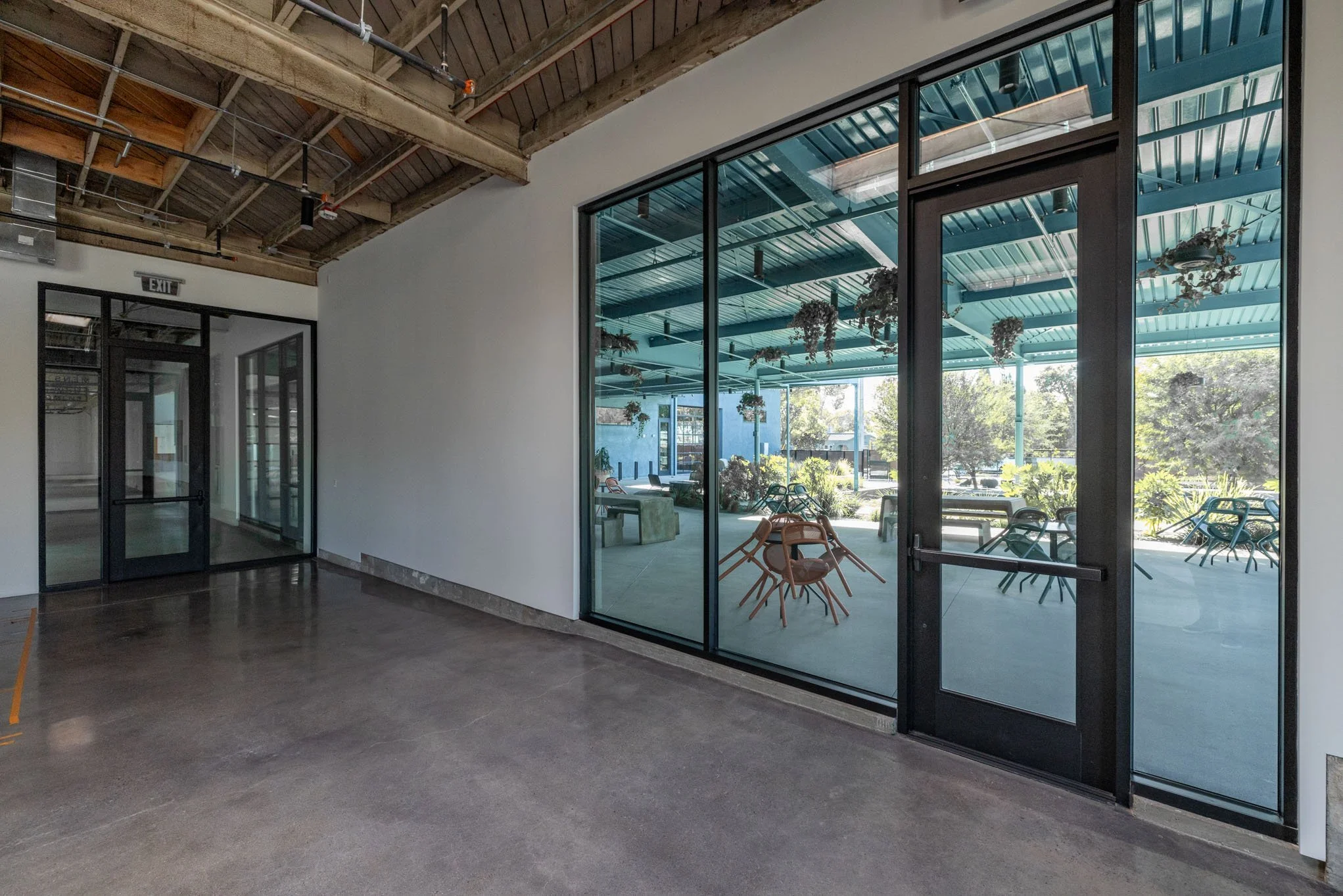 Interior view of a modern building with large glass doors and windows, showing a covered outdoor seating area with chairs and hanging plants, bright natural light, and trees outside.