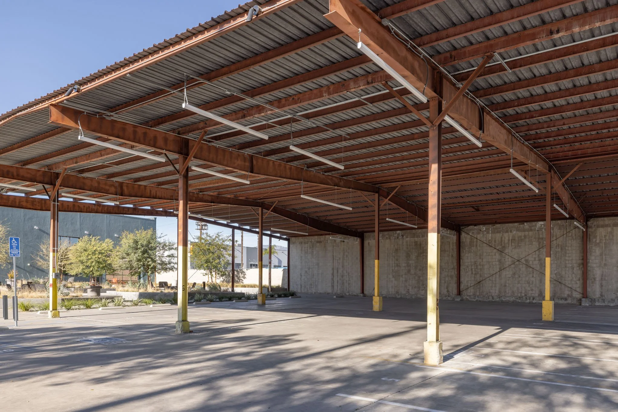 Empty parking lot with a large metal awning, yellow parking space dividers, and trees in the background.