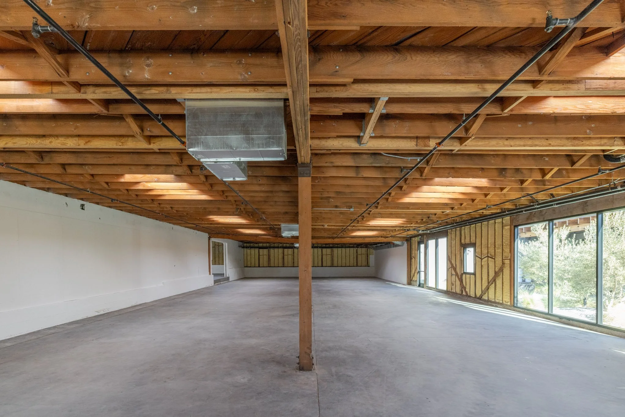 Empty commercial space under construction with wooden ceiling, large windows on the right, concrete floor, and exposed HVAC ductwork