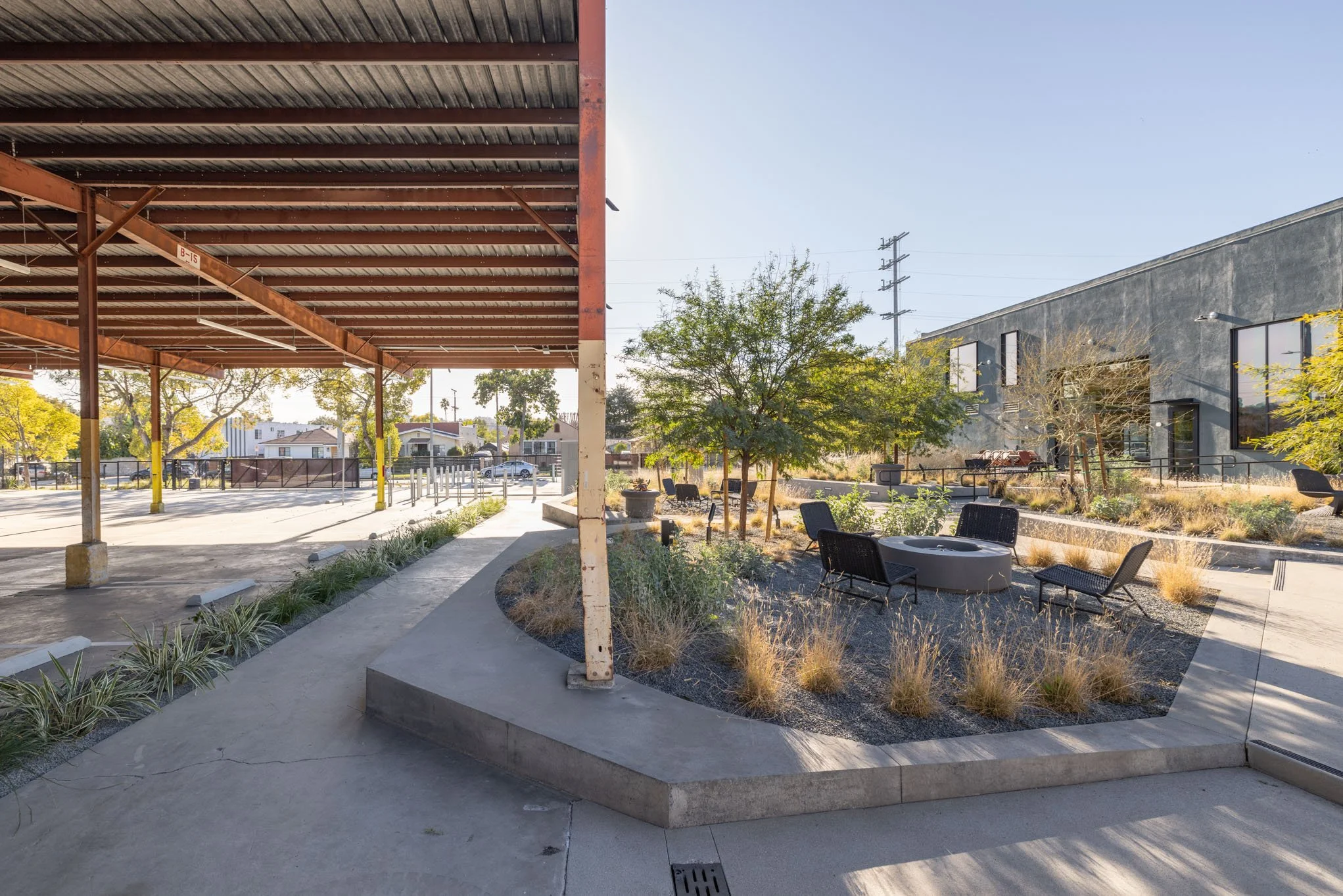 An outdoor urban area with a shaded structure on the left, a landscaped section with chairs and a fire pit in the center, and a modern building with large windows on the right. The scene features trees, plants, and street in the background.