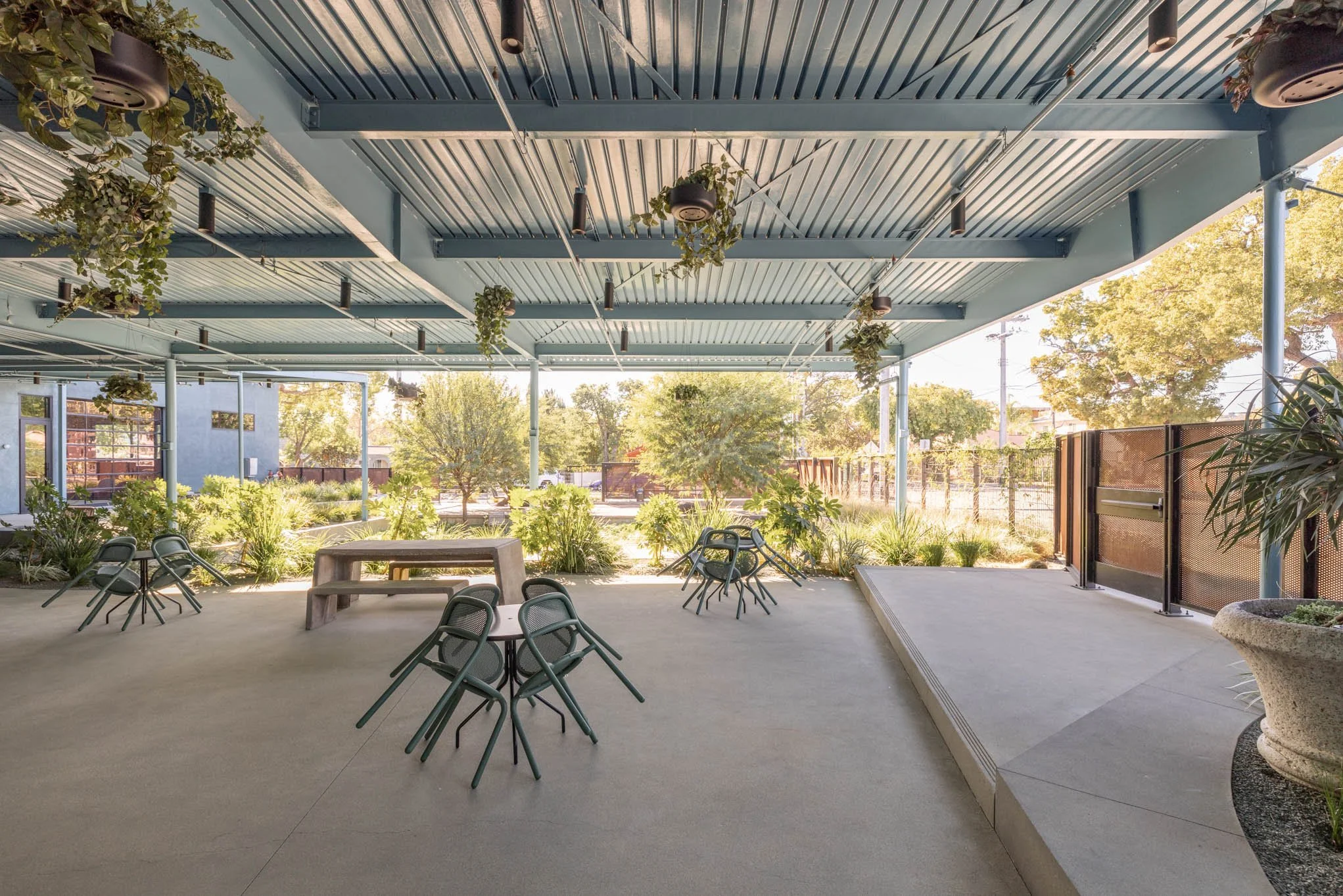 Open patio with metal chairs, potted plants, and greenery under a blue ceiling, sunlit with trees and a building in the background.