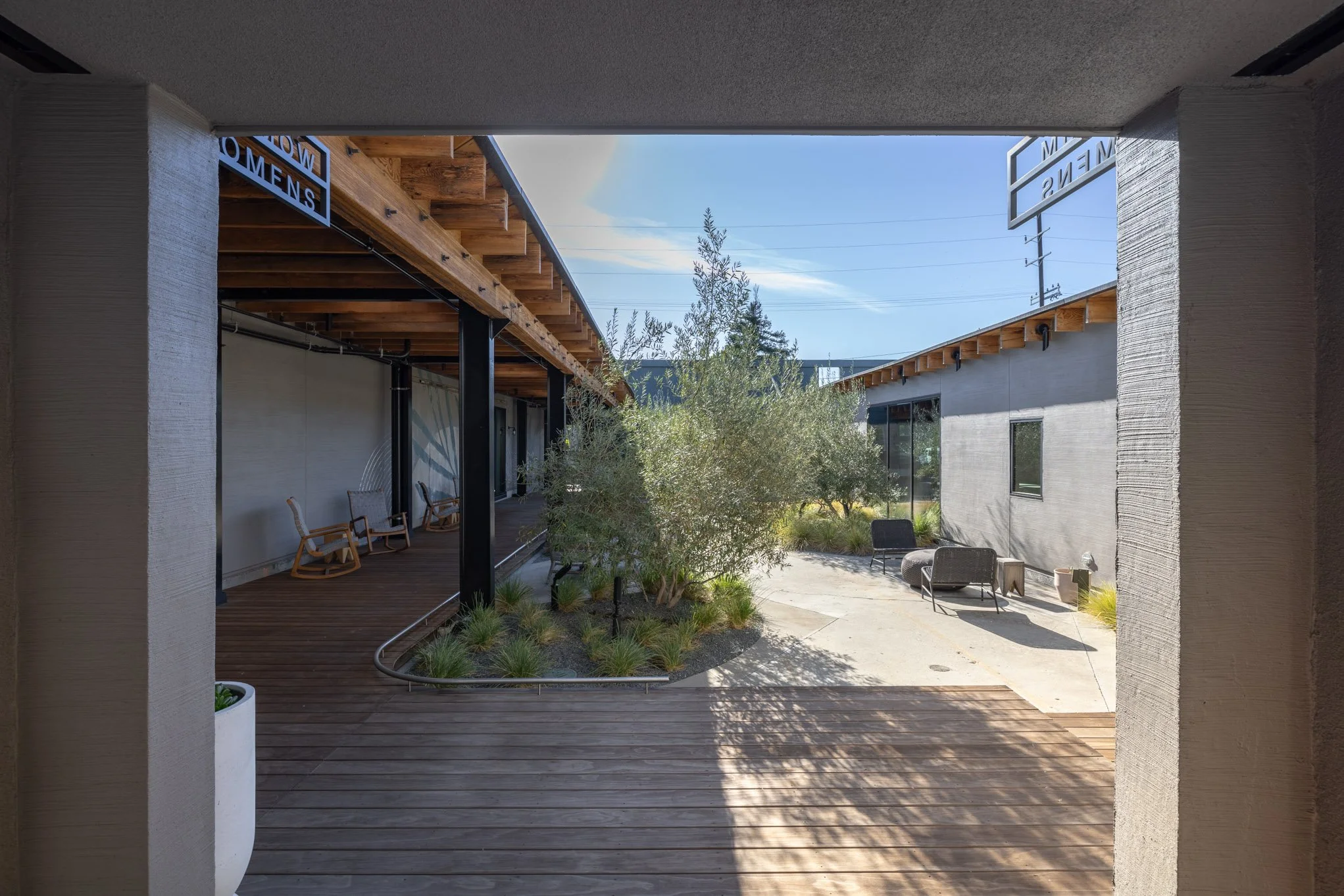 A modern outdoor courtyard with a central tree, seating furniture, and a mix of wooden and concrete surfaces, viewed from under a covered walkway.