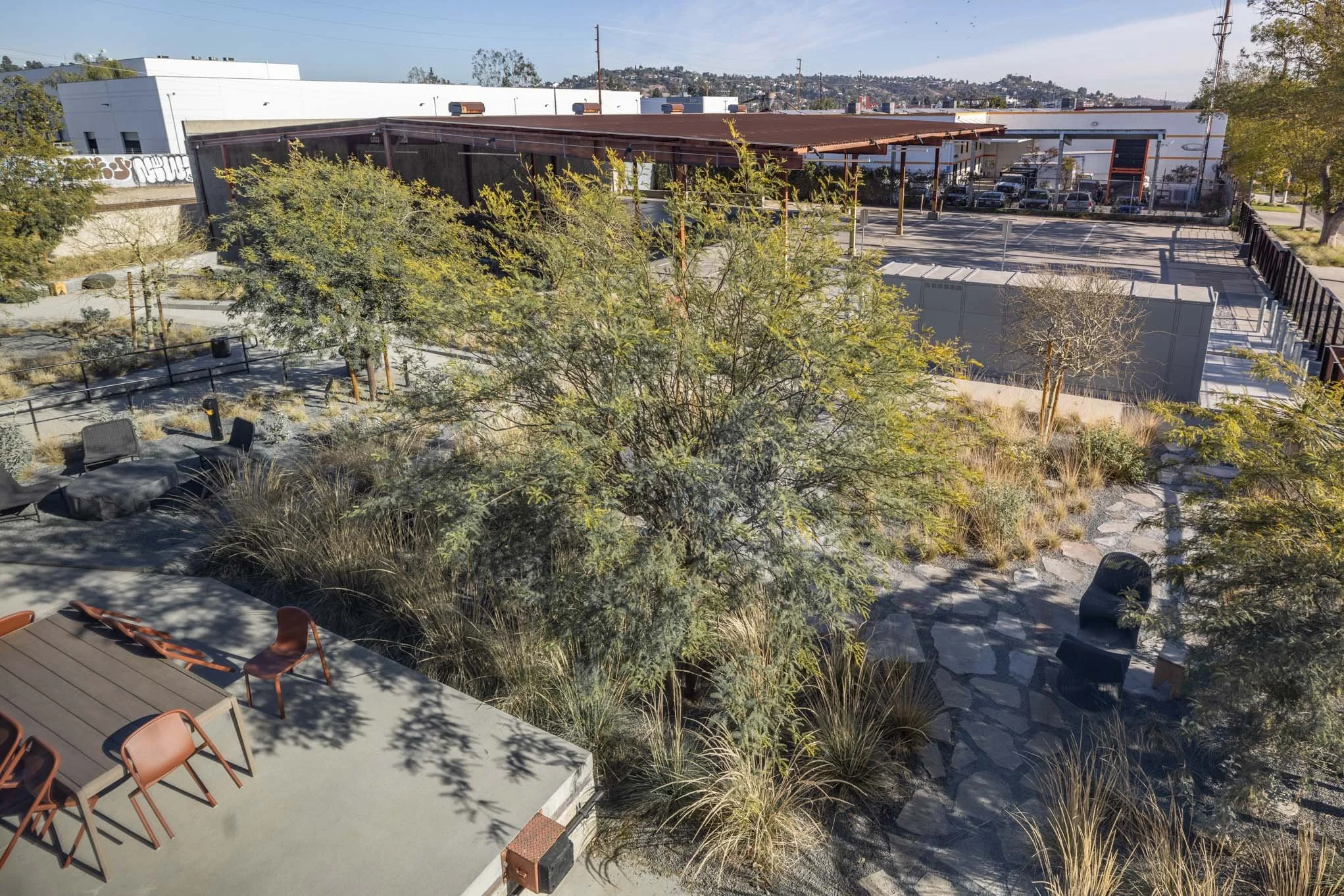 Rooftop garden with seating area, trees, and plants. Urban environment with parking lot and industrial buildings in the background.