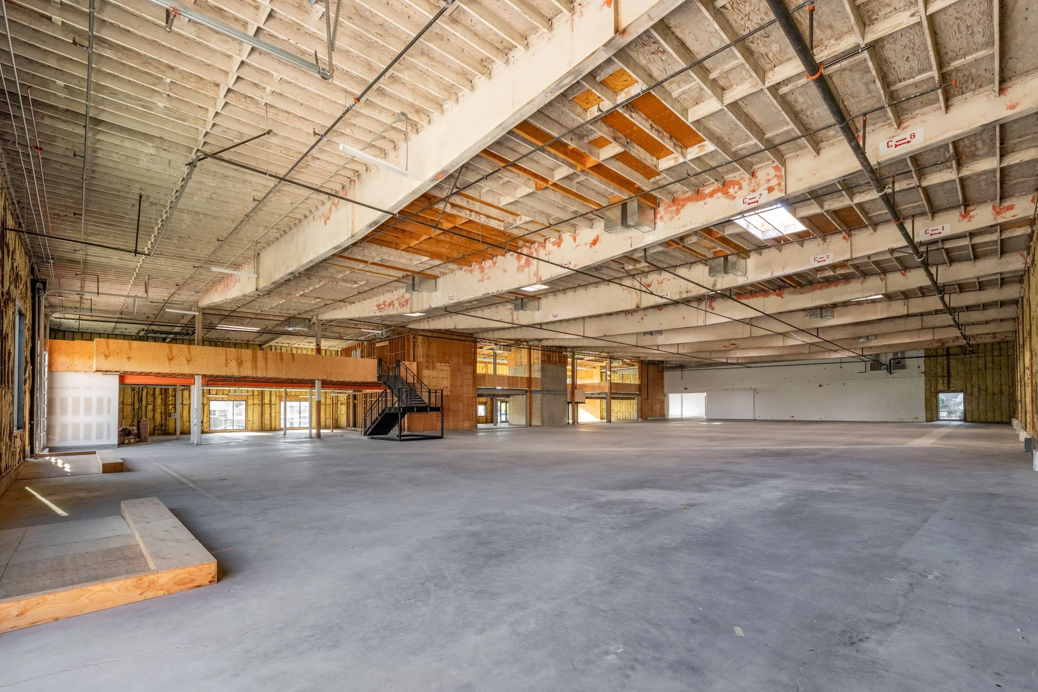 Large open unfinished building with exposed ceiling beams, walls wrapped in insulation, and a concrete floor. There is a black staircase leading to an upper level and several window openings along the walls.