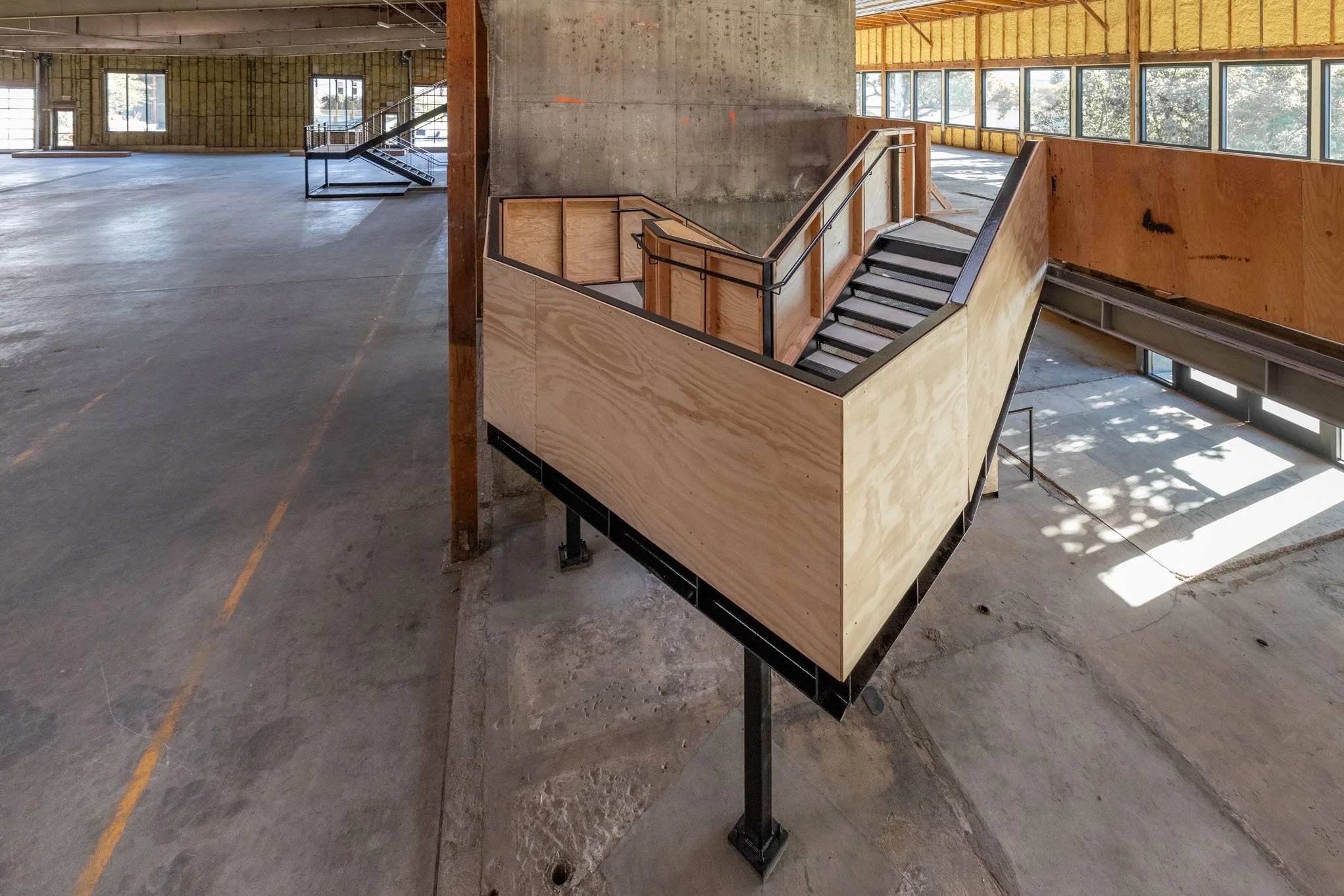 Construction site of a building with staircases and unfinished walls, featuring large windows and exposed concrete and wood framing.