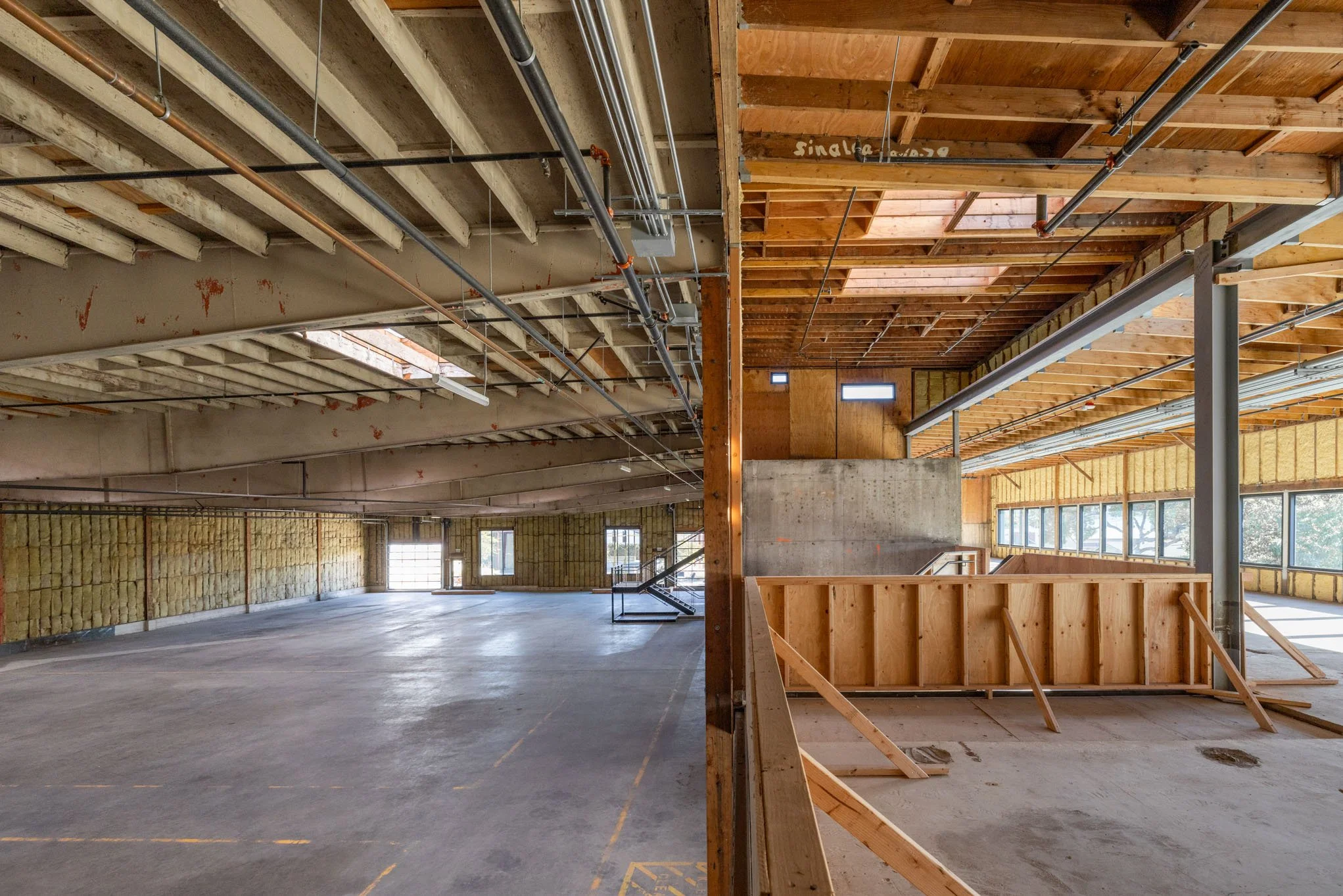 Split view of two construction sites: the left shows an unfinished building with exposed ceiling beams, pipes, and a concrete floor, while the right depicts a partly finished wooden structure with windows, insulation, and a staircase.