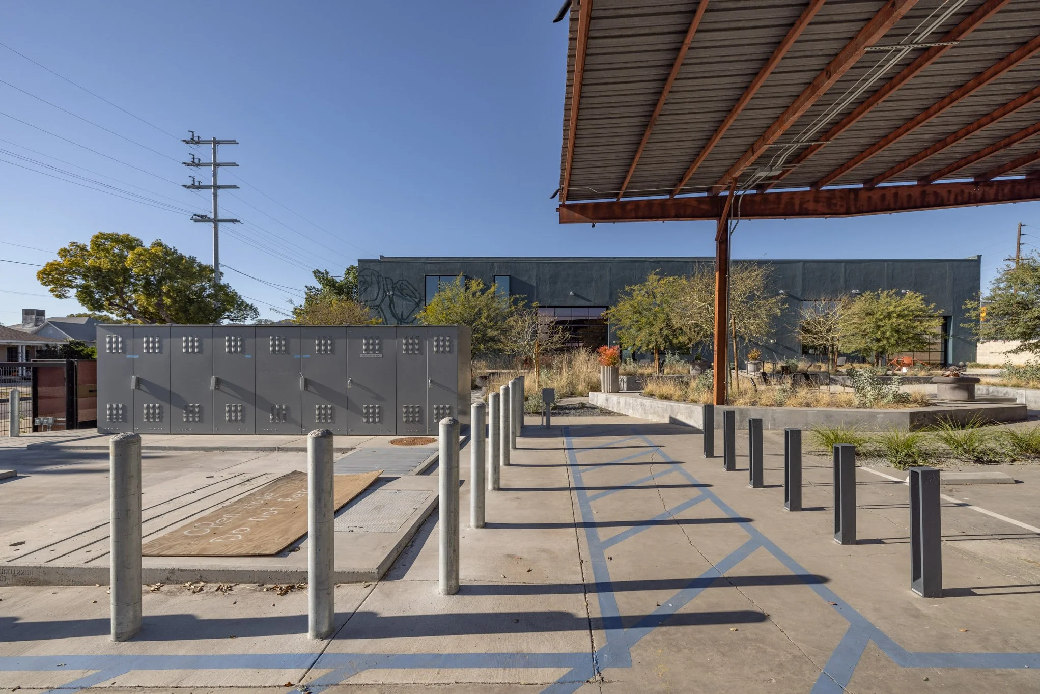 Empty outdoor area with bike racks, metal lockers, trees, and a shaded structure, under clear blue sky.