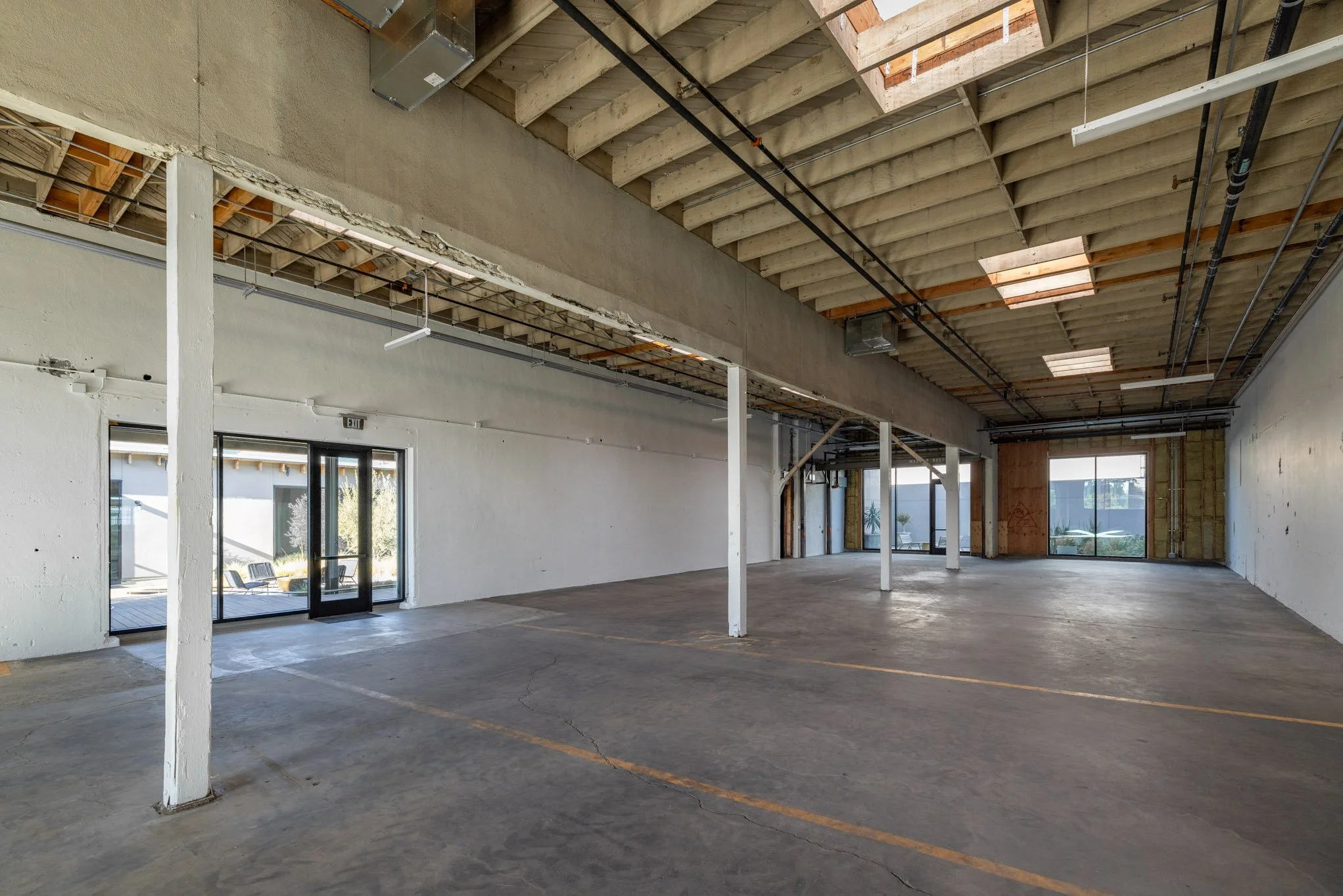 Empty commercial interior space under construction with concrete floors, white walls, exposed ceiling with wooden beams and skylights, glass doors, and windows.