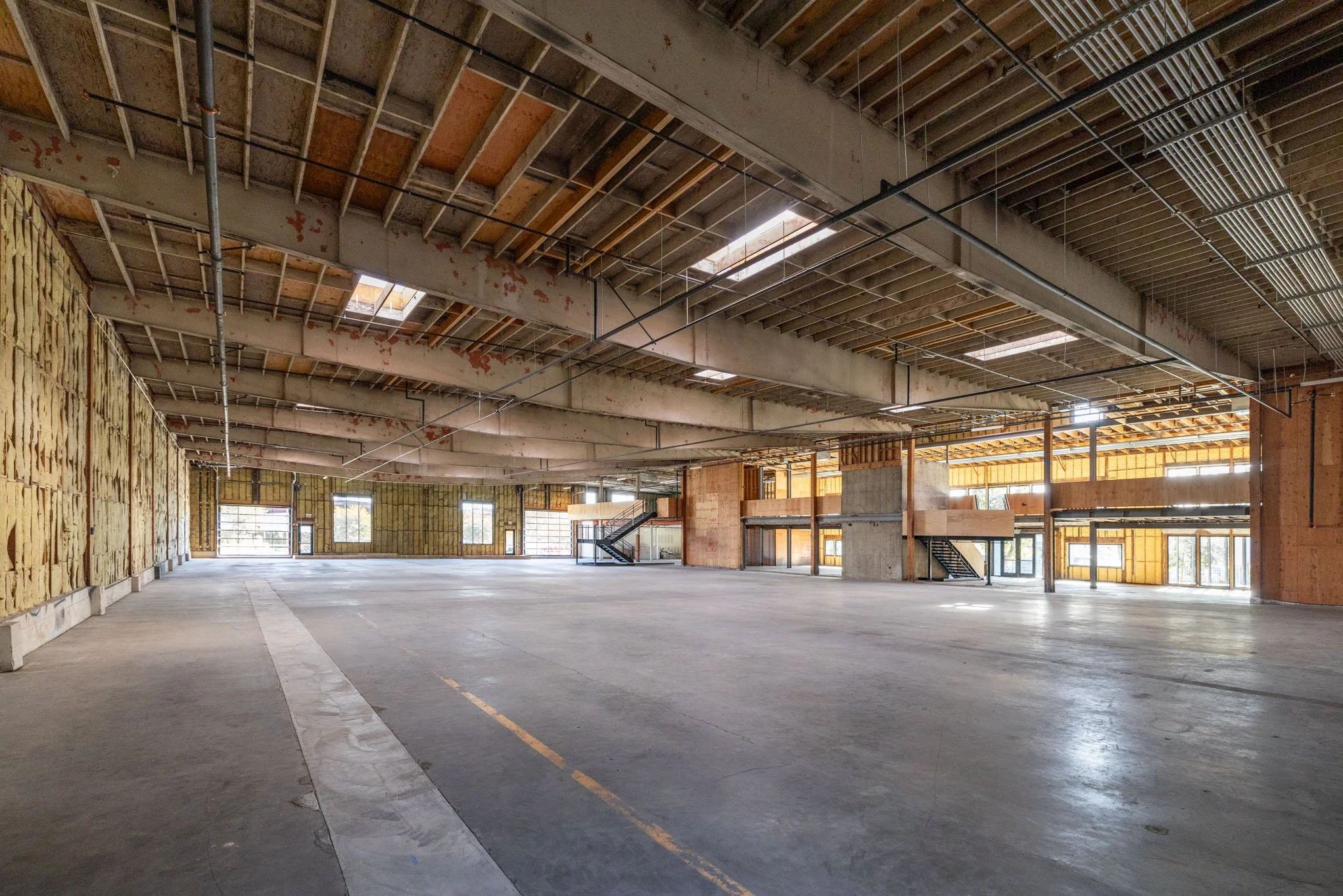 Empty interior of a building under construction with exposed ceiling beams, insulation on walls, and large windows.