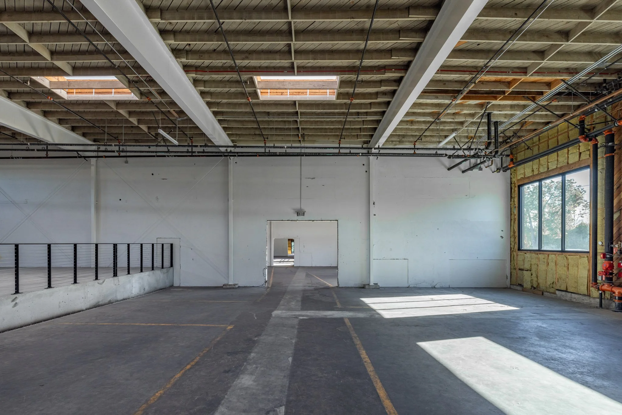 Empty indoor parking garage with a concrete floor, unfinished walls, large window, and exposed ceiling with ducts and pipes.