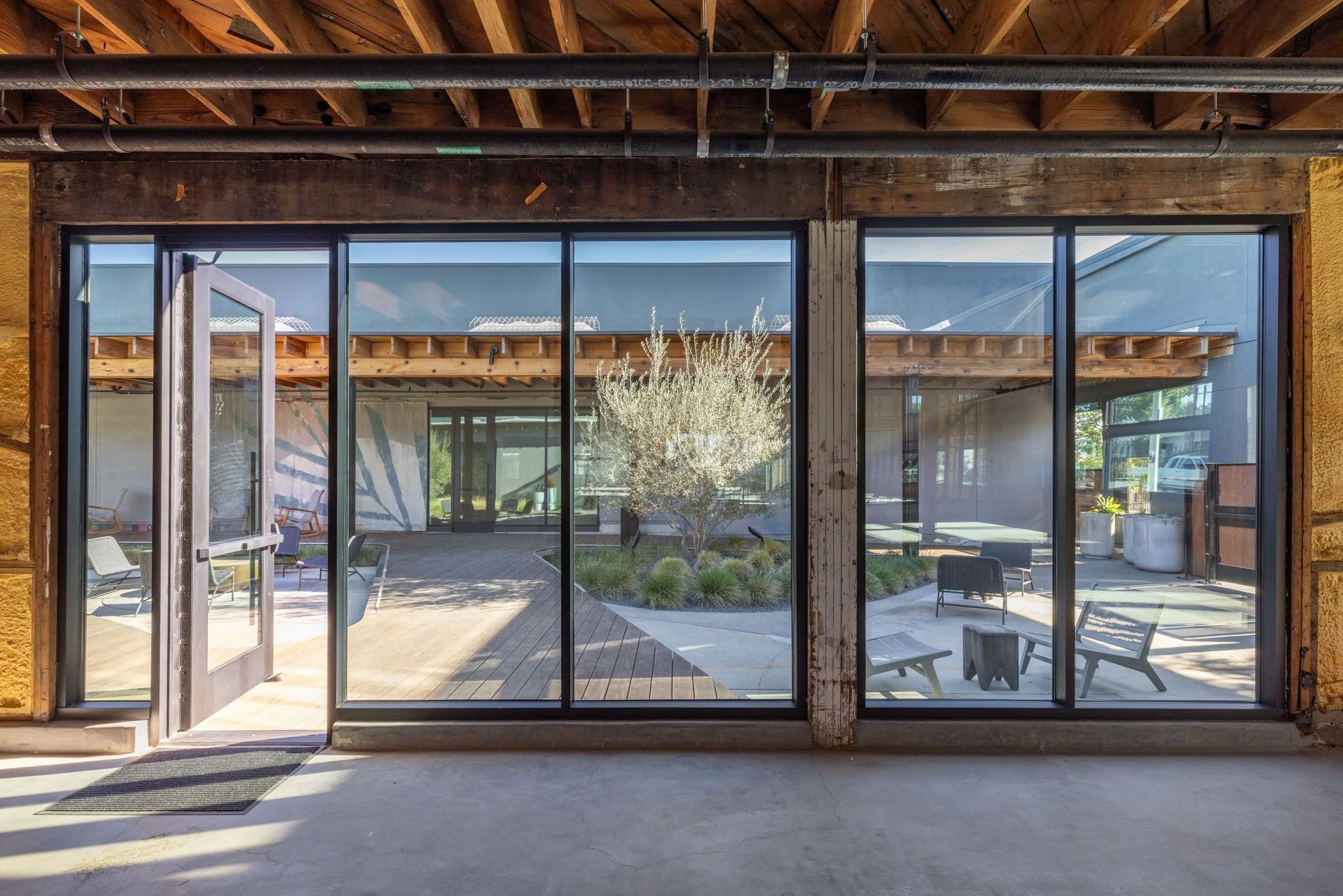View through large glass sliding doors into an outdoor patio with chairs, a tree, and landscaping.