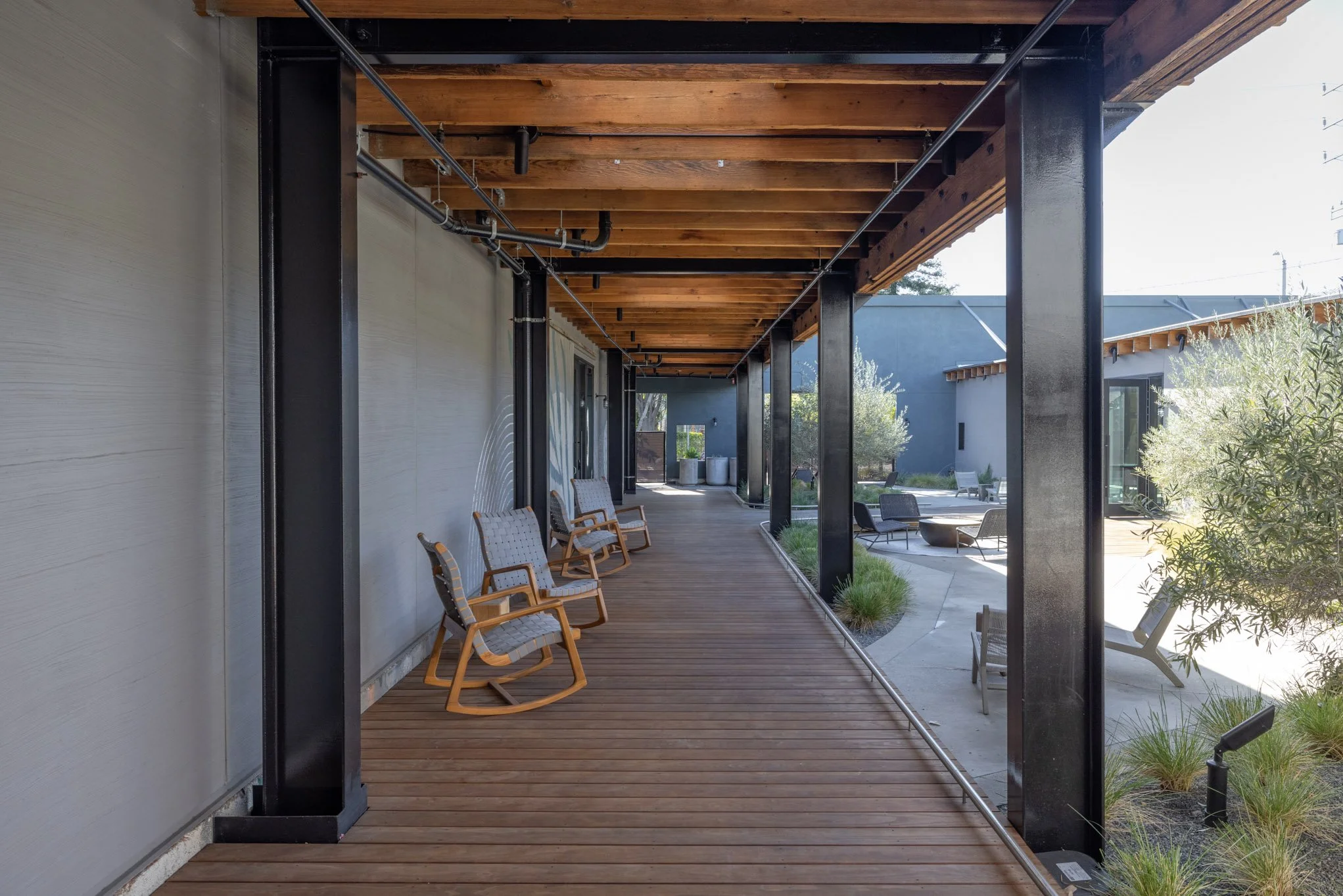 Modern outdoor patio with wooden flooring, black metal columns, and a wooden beam ceiling, featuring chairs along the covered walkway and seating areas with tables and greenery in the background.