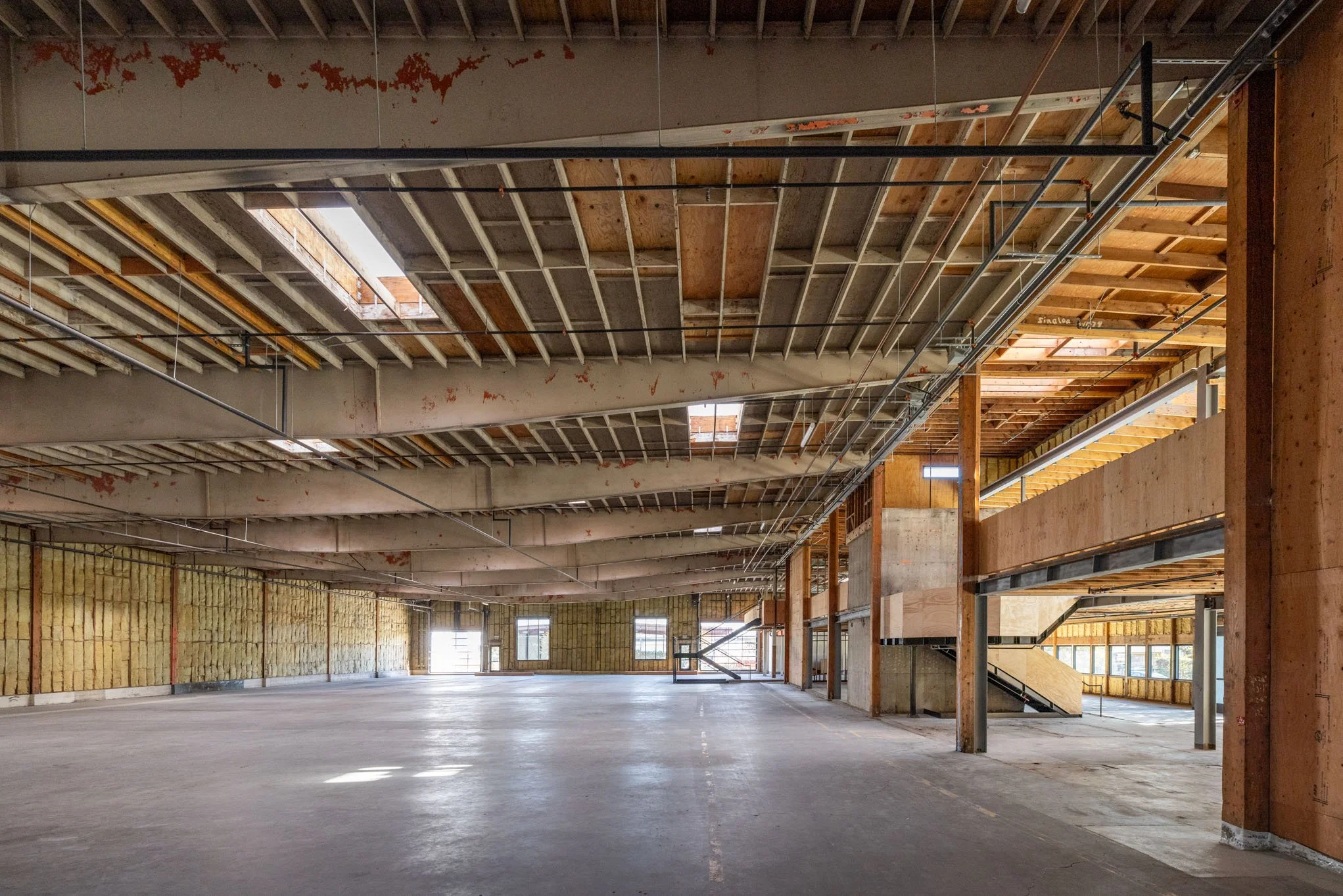 Interior view of a building under construction with exposed wooden framing, insulation, metal pipes, and no finished walls or ceiling.