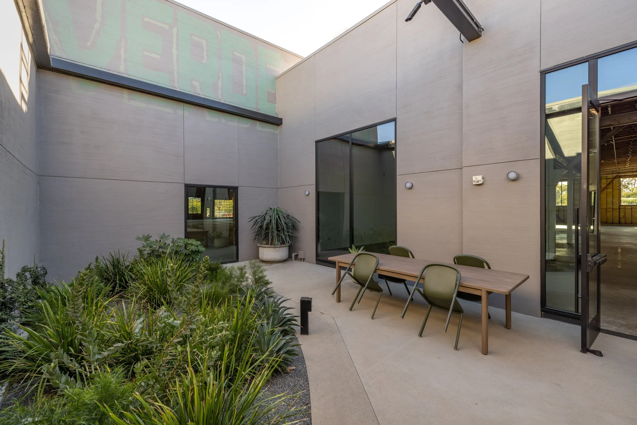 Outdoor patio with table, chairs, and greenery, part of a modern building with large windows and beige exterior walls.