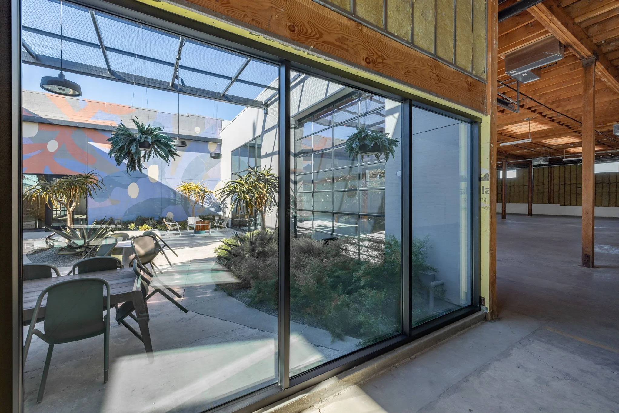 View through glass doors into a courtyard with tropical plants and trees, chairs, and a colorful mural on the wall.