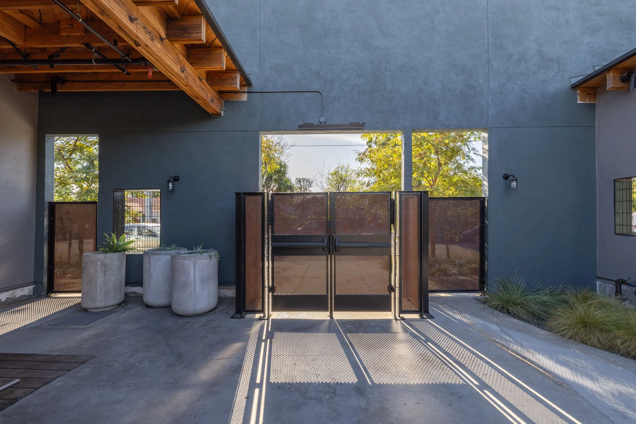 Modern outdoor entrance with black metal gate, concrete planters with succulents, and trees visible through the openings in the wall.