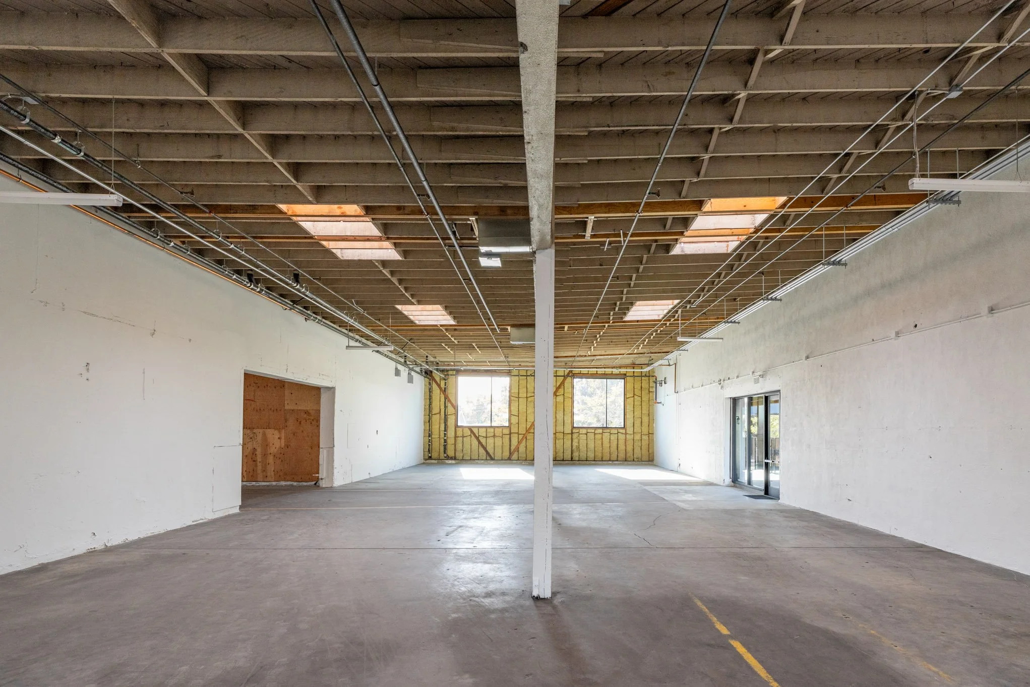 Empty interior of a building under construction with unfinished walls, exposed ceiling with wooden beams and lots of electrical wiring, and windows at the back allowing natural light.