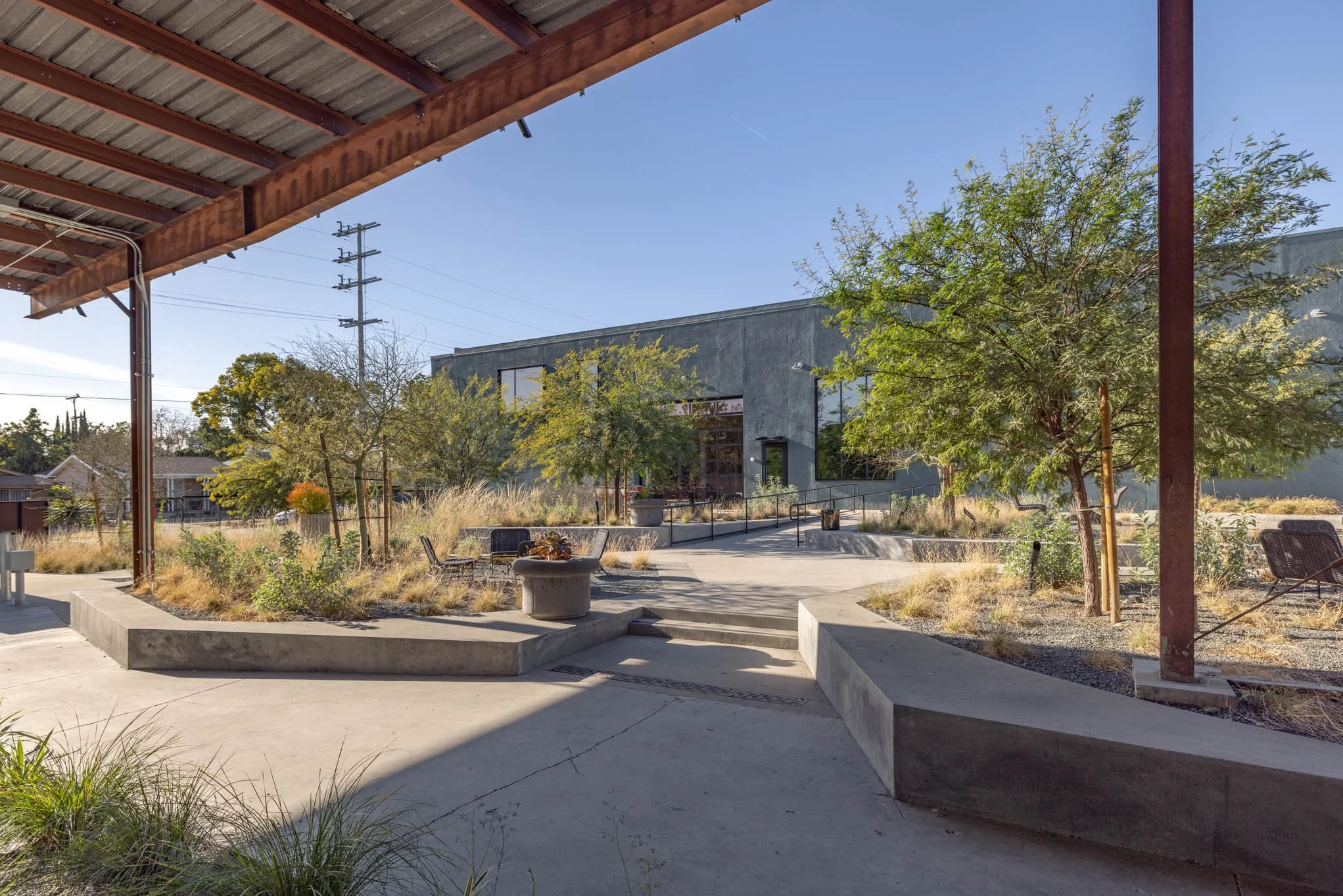 Outdoor patio area with concrete seating, trees, and a modern building in the background under a clear blue sky.