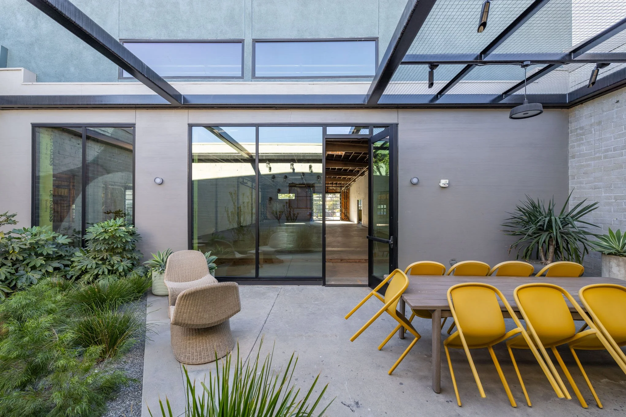 Outdoor patio with yellow chairs around a wooden table, wicker chairs, green plants, and glass sliding doors leading indoors.